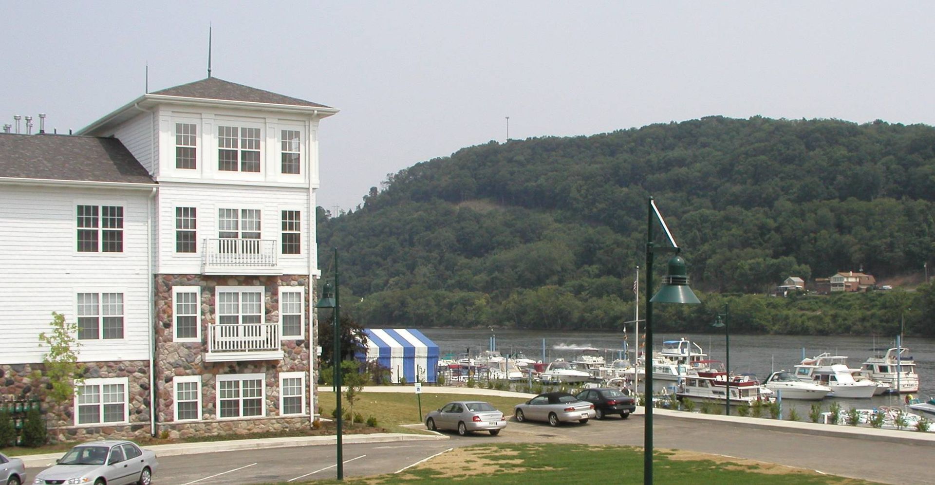 Harbor with parked cars, white building, and green hillside under a cloudy sky
