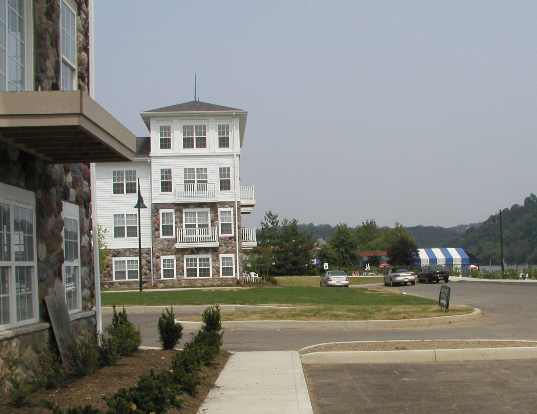 White multi-story building by a road with lawn, trees, and blue-striped tents in the distance.