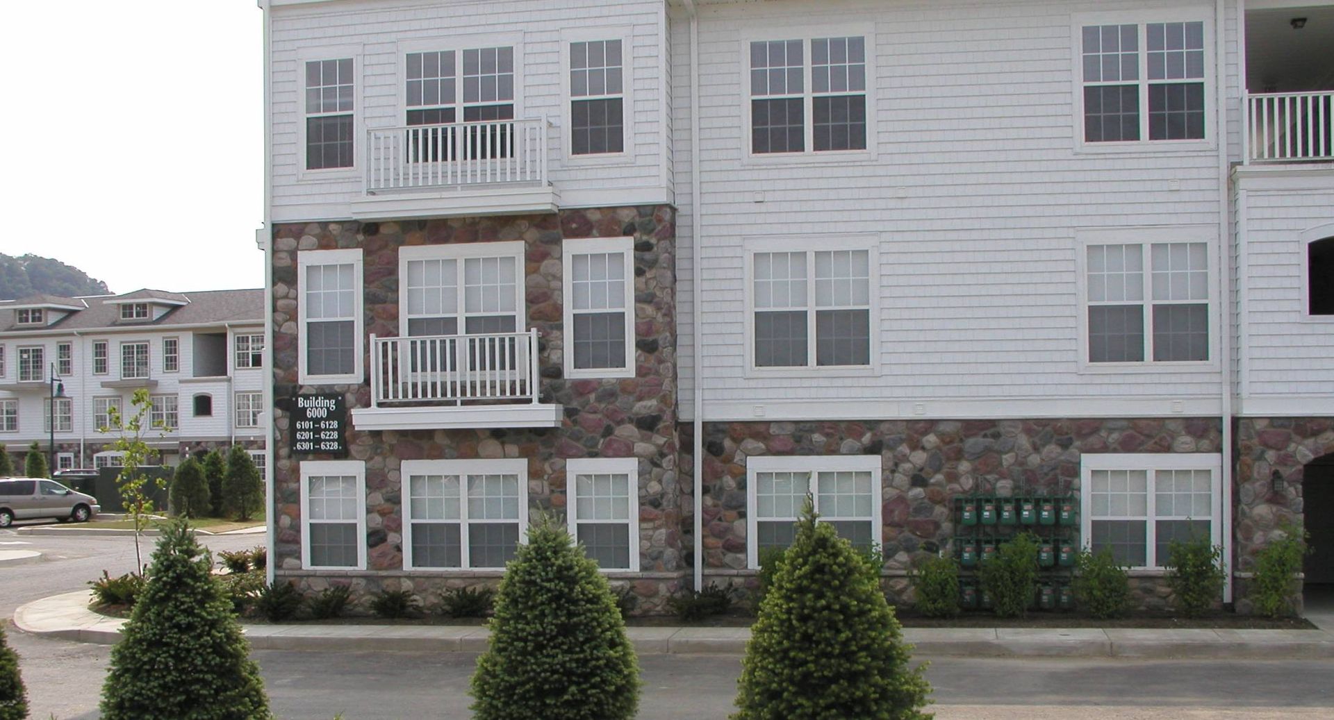 Apartment building exterior with white siding, stone base, and small balconies behind neatly trimmed shrubs