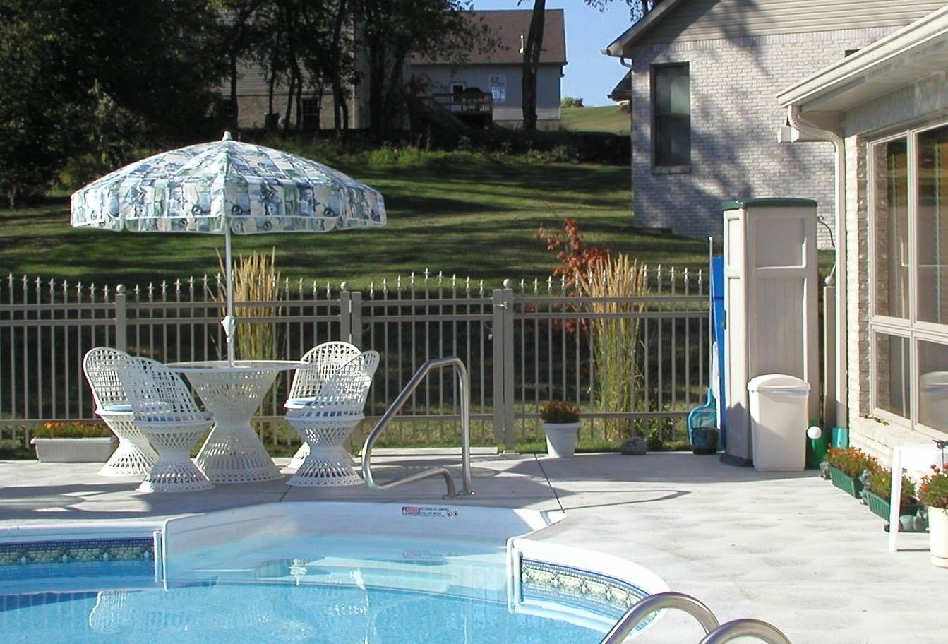 Backyard pool patio with white chairs, umbrella, fence, and a house in the background