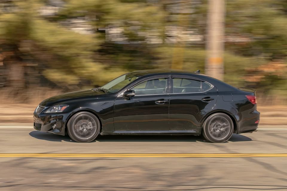 A black car is driving down a road with trees in the background.