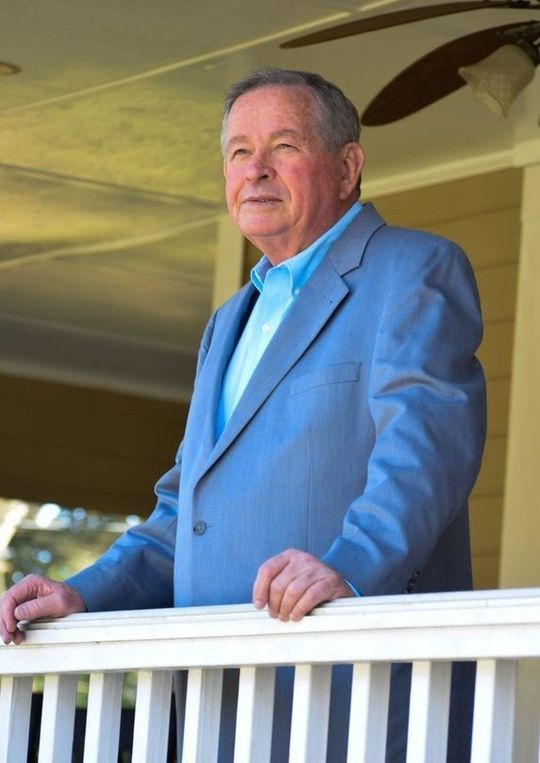 Man in a blue suit standing on a porch, leaning on a white railing and looking outward.