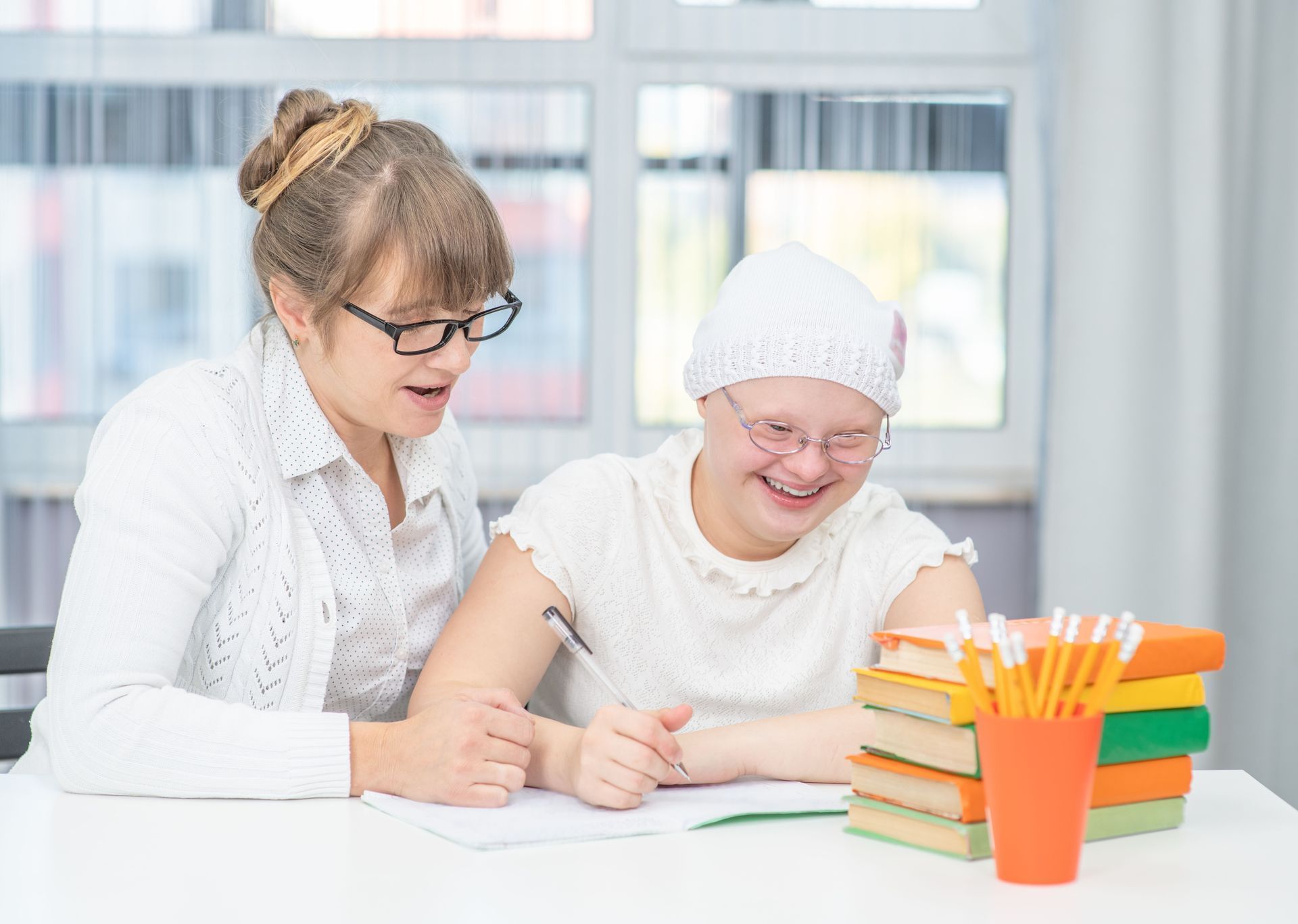 Teacher helping a smiling student write at a desk with books and pencils in a bright classroom