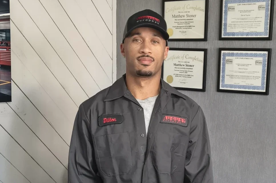 Portrait of a mechanic standing indoors in front of framed certificates on a gray wall.