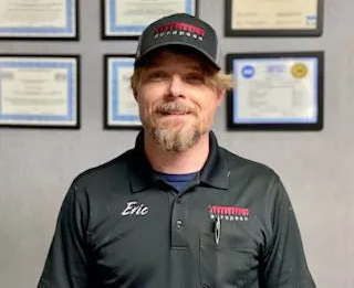 Man in black cap and polo shirt standing indoors before framed certificates and awards.
