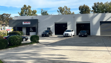 Gray auto repair shop with three car bays and parked vehicles in front under sunny skies