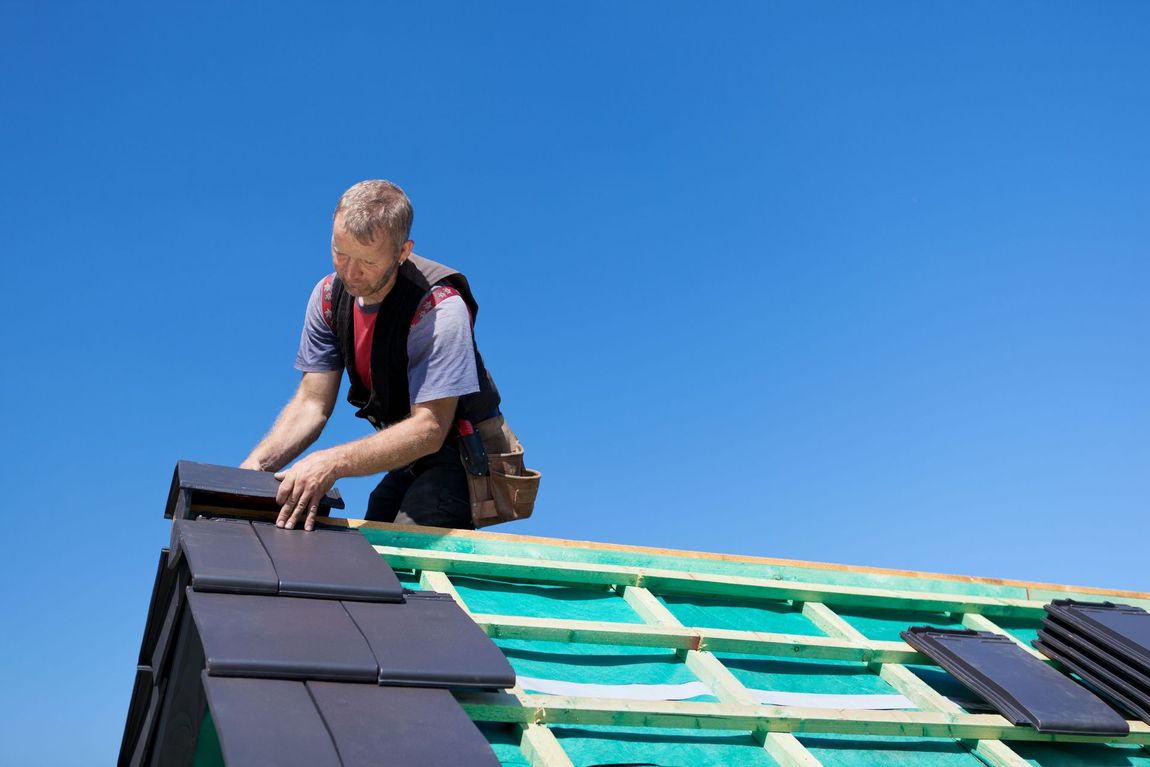 Roofer installing black roof tiles on a pitched residential roof under clear blue skies, using safety gear and working on a green underlayment and wooden battens.