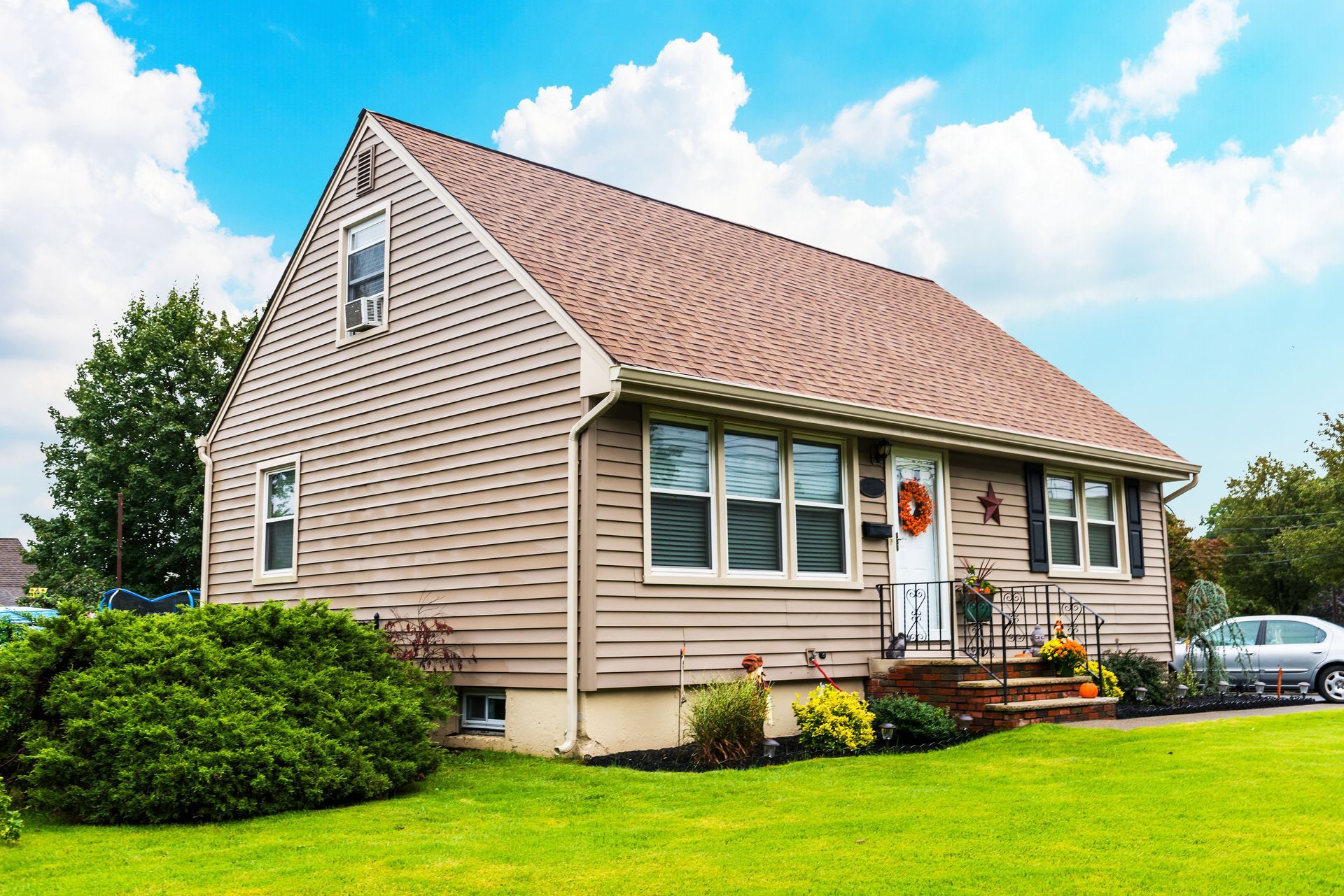 Single-story suburban house with beige vinyl siding, a brown shingle roof, and a well-maintained front yard featuring green grass, shrubs, and seasonal decorations on the porch.