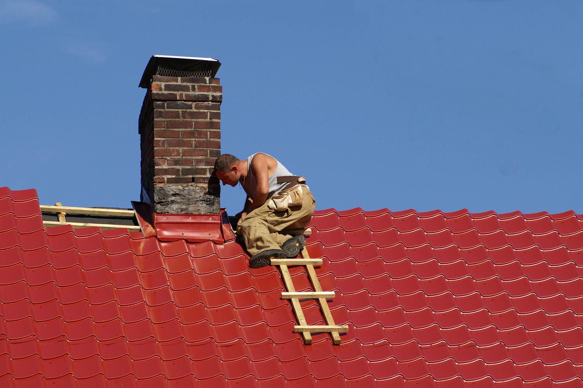 Roofer working on red metal roofing around a brick chimney, using a small ladder for support under a clear blue sky.