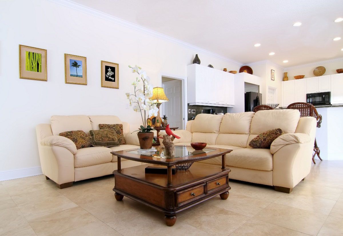 Bright and modern living room with beige leather sofas, a wooden coffee table with glass top, decorative pillows, and an open kitchen in the background with white cabinetry and bar stools.