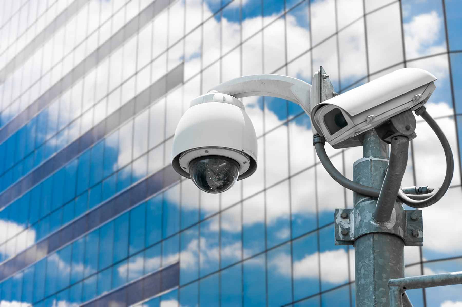 Two surveillance cameras mounted on a pole against a glass office building and blue sky