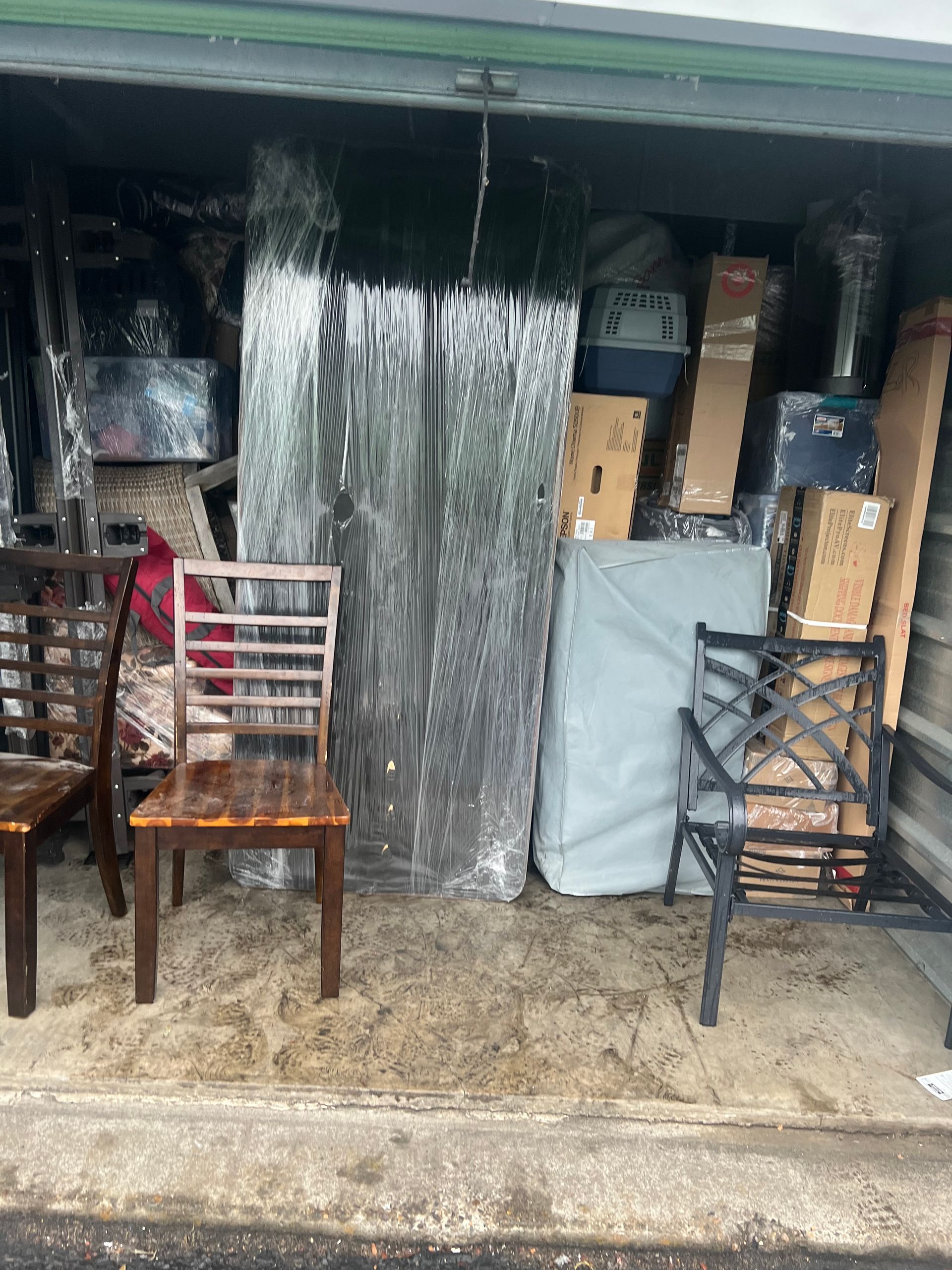 A storage unit filled with furniture and boxes, including chairs and a wrapped mattress.