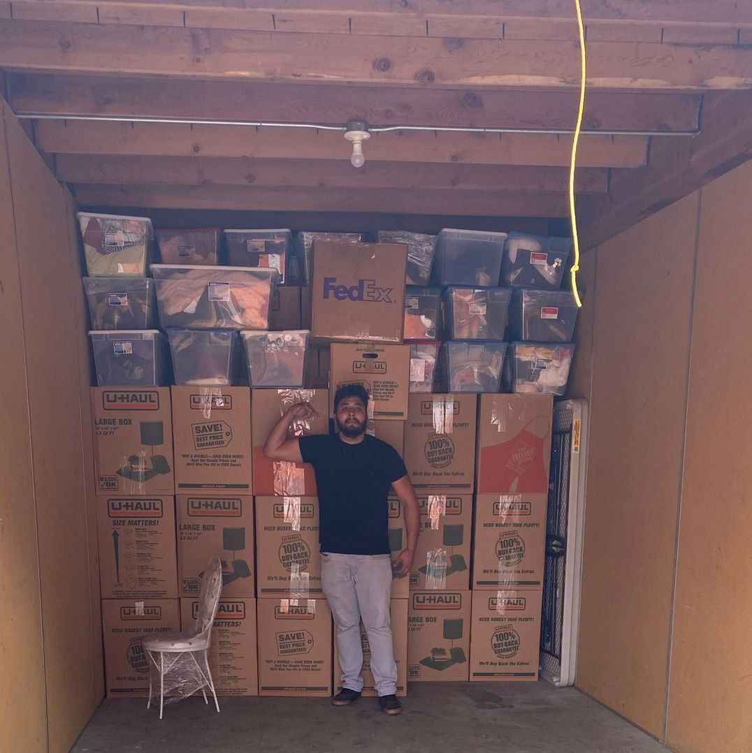 Man standing in storage unit filled with boxes; raising arm. Brown boxes and bins stacked up, small chair to the left.