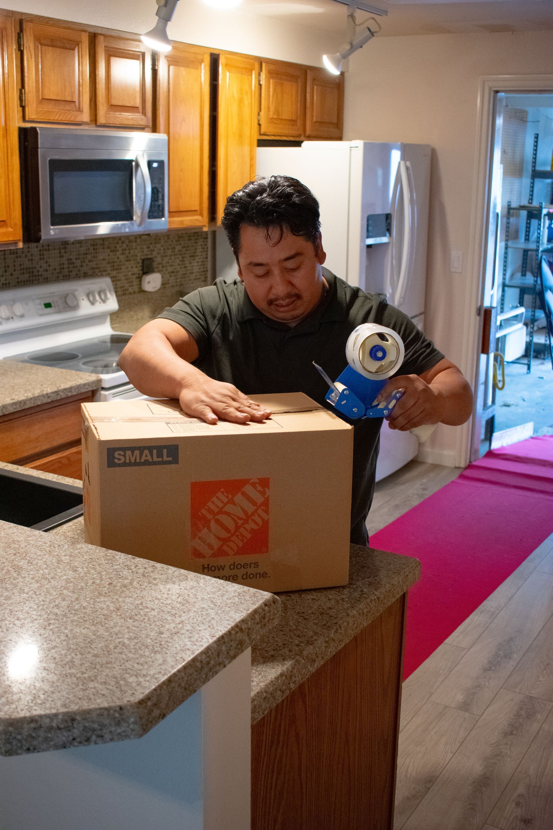 Man taping a cardboard box in a kitchen. The box is on a countertop, near the stove and refrigerator.