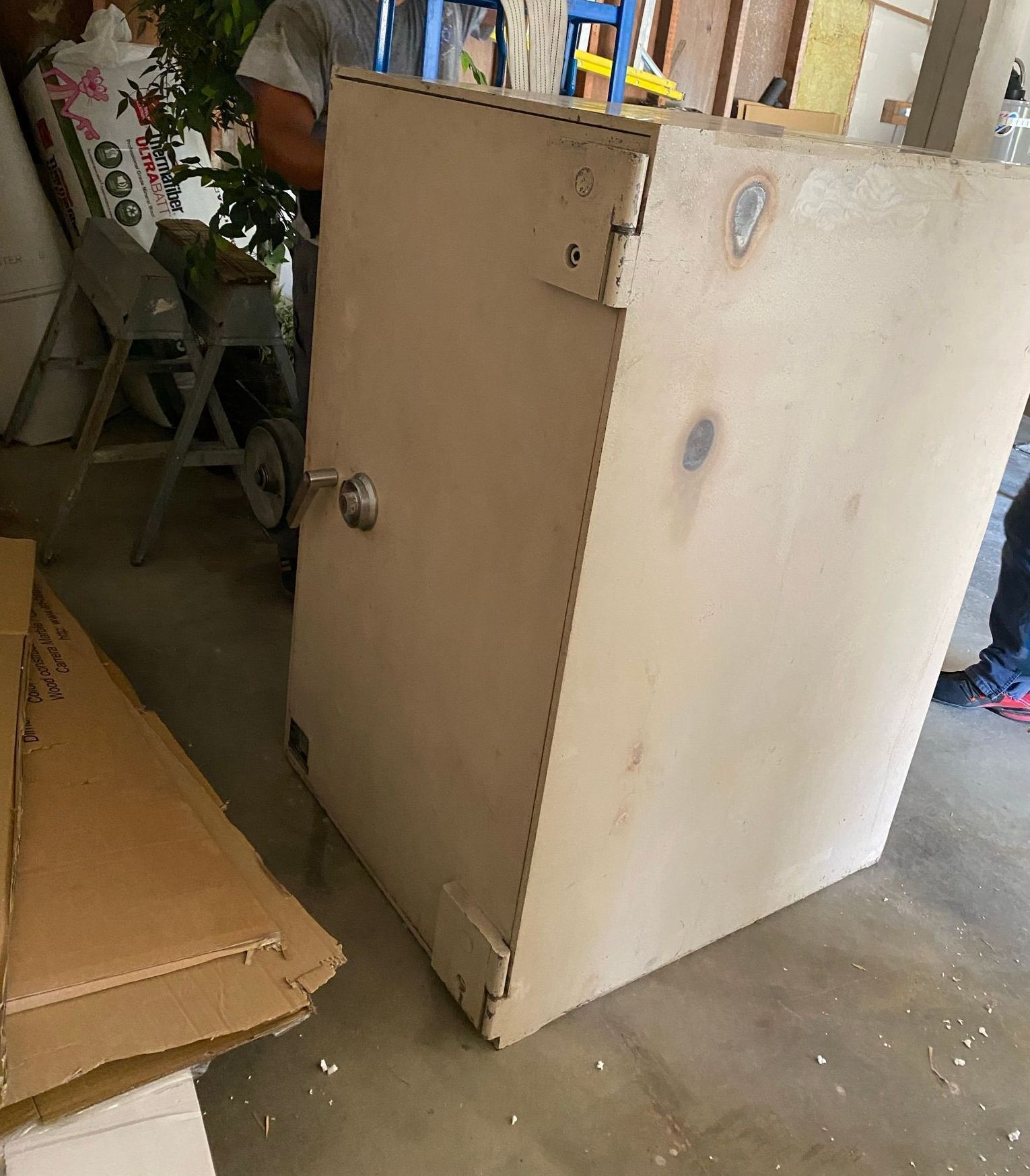 A large, beige safe with a silver handle sits on a concrete floor near cardboard and a person.
