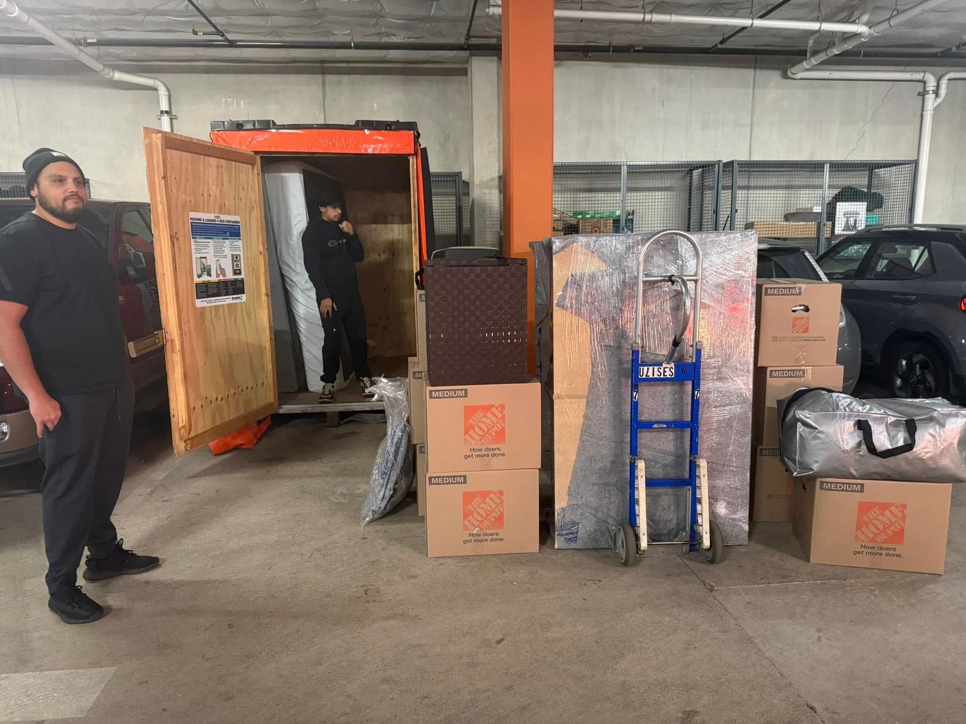 Two people loading a truck with boxes and furniture in a parking garage. One person looks at the camera.
