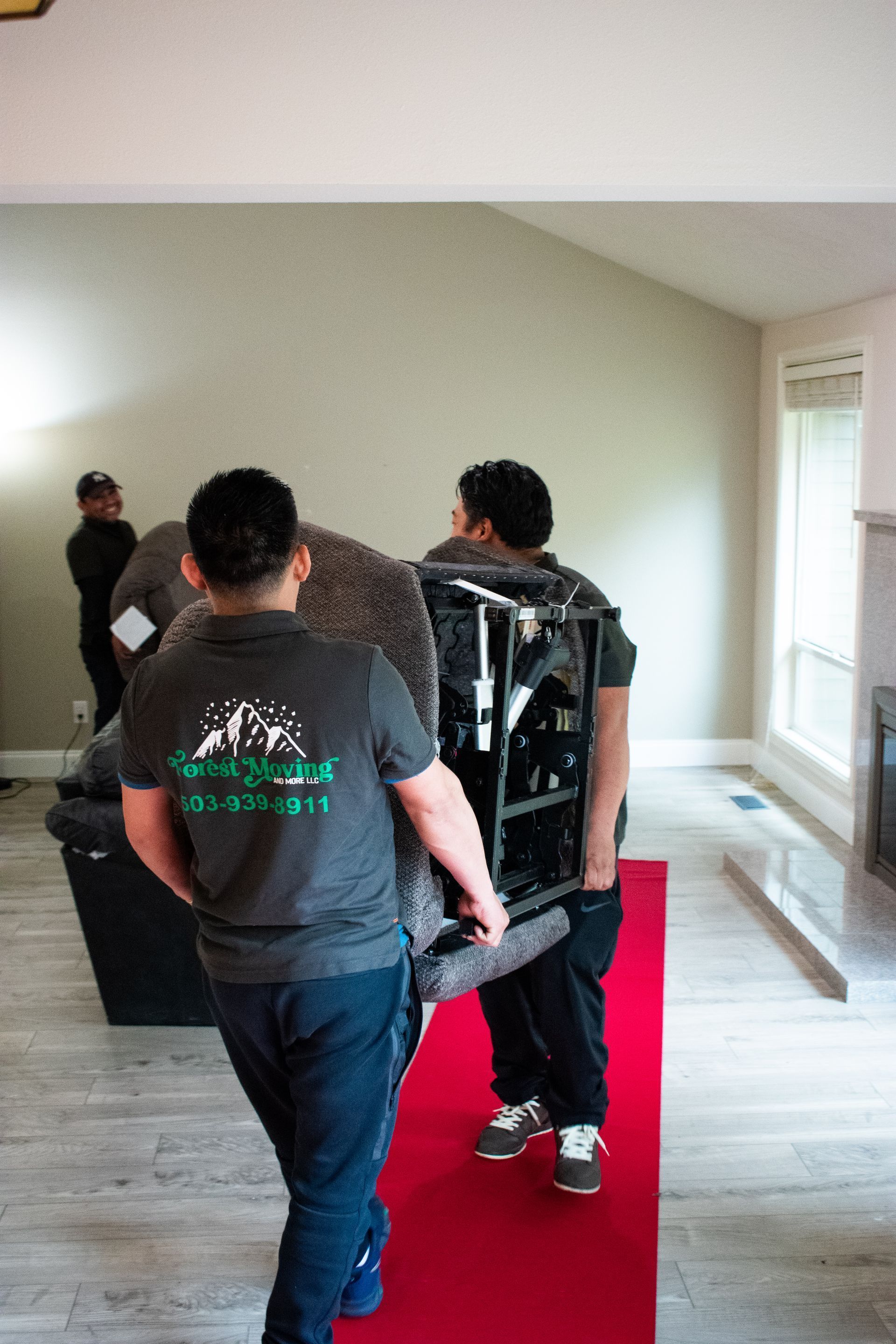 Two movers carrying furniture on a red carpet in a room with a third person observing.