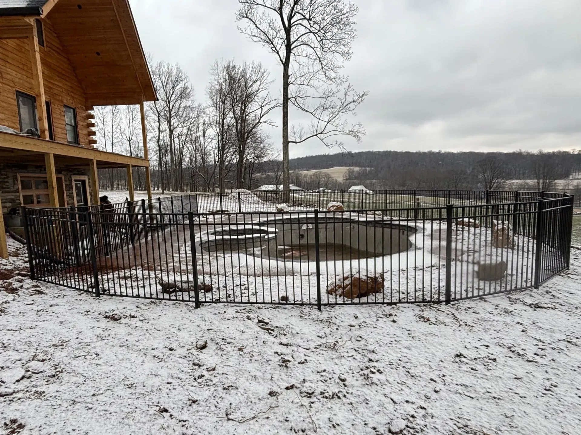 A swimming pool surrounded by snow and a fence in front of a log cabin.