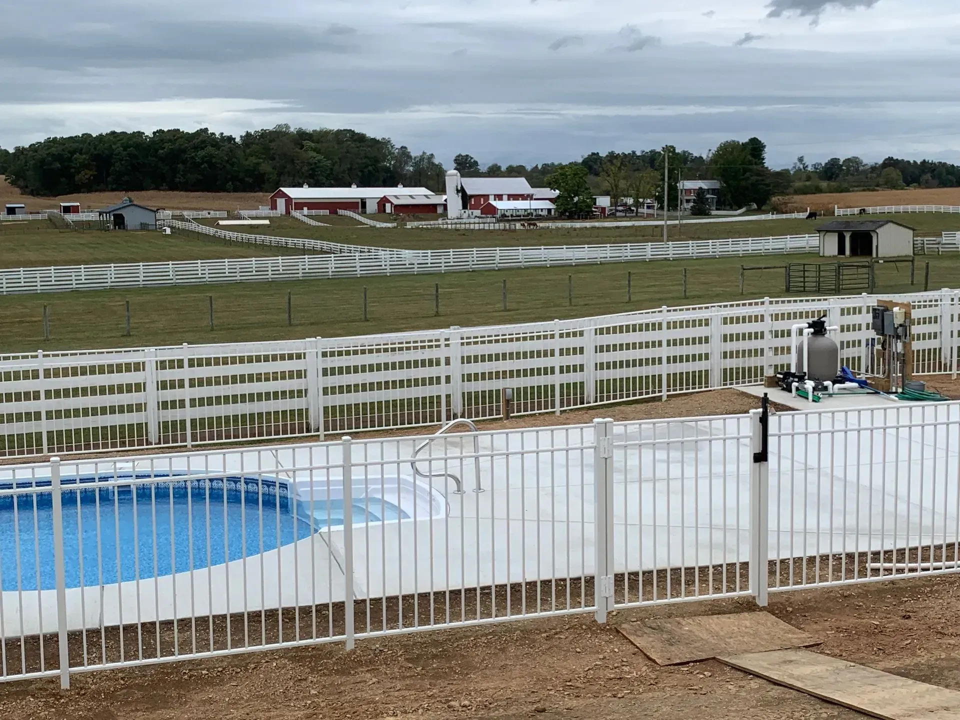A white fence surrounds a swimming pool with a farm in the background.