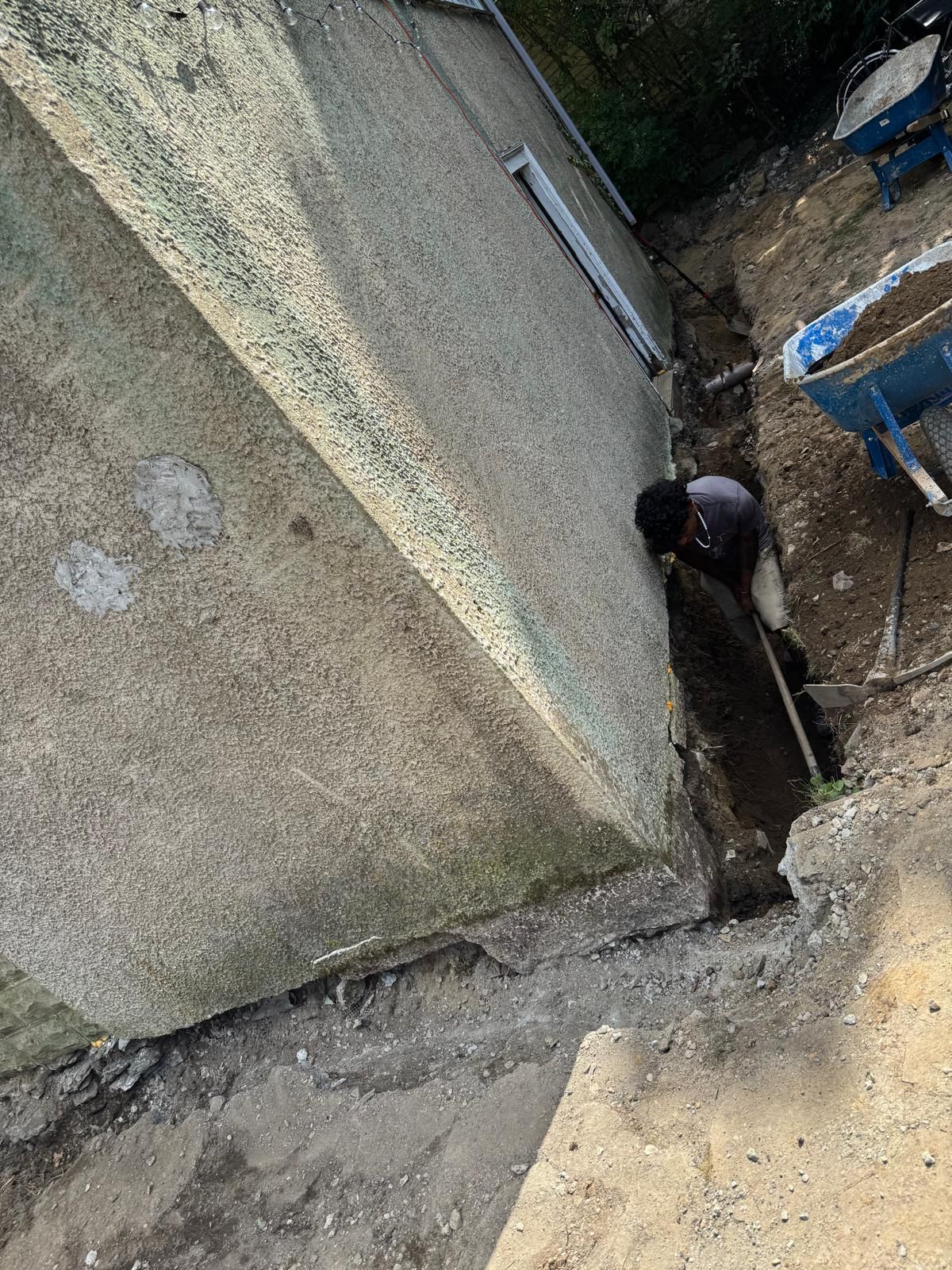 A worker digs a narrow trench alongside the exterior wall of a building, with two wheelbarrows nearby.