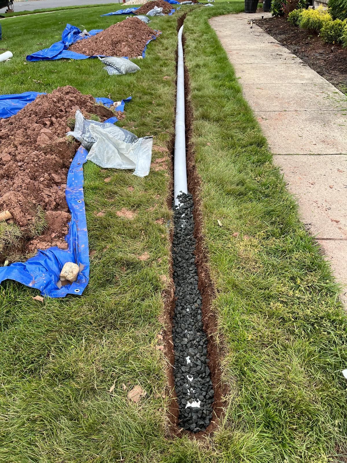 A long trench in a grass lawn, featuring a white pipe partially covered by gravel, with piles of dirt on blue tarps nearby.
