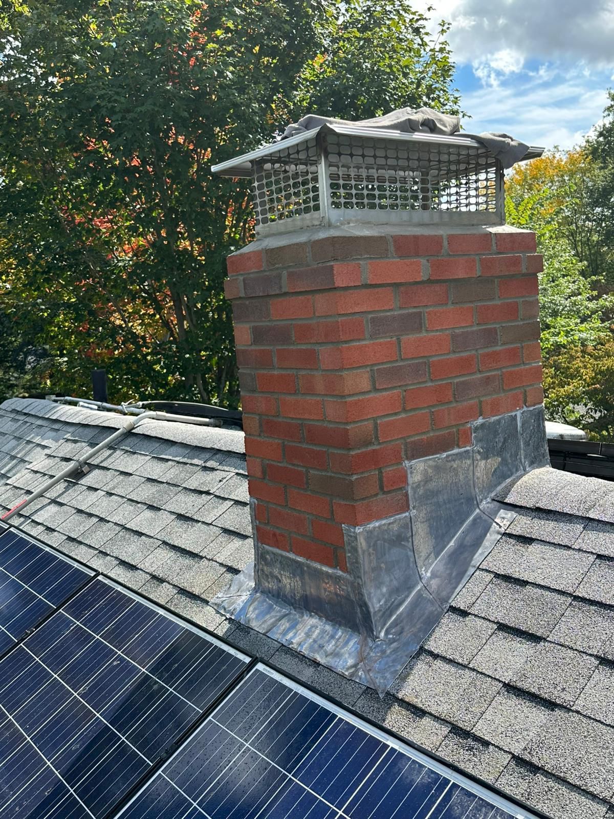 A brick chimney with a metal cap and lead flashing sits on a residential roof next to solar panels.