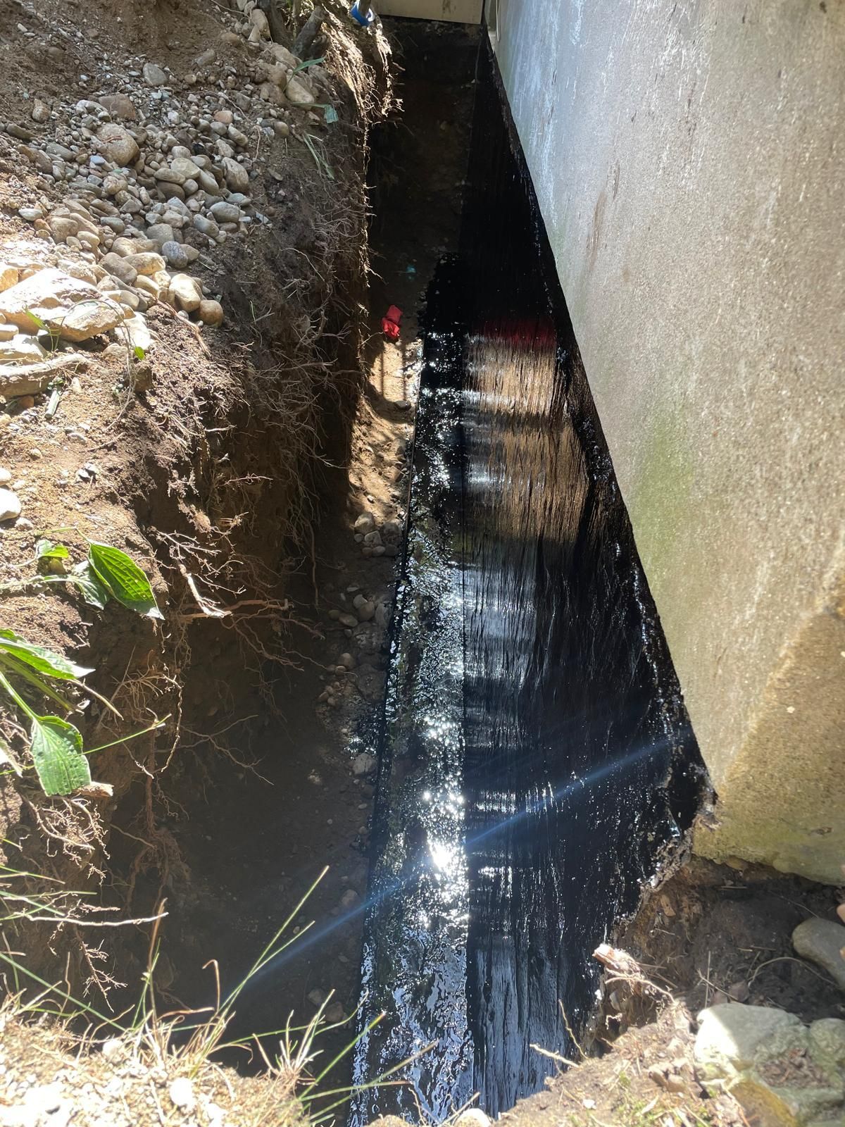 A narrow trench dug beside a concrete foundation wall, which is coated in a thick, black waterproof sealant.