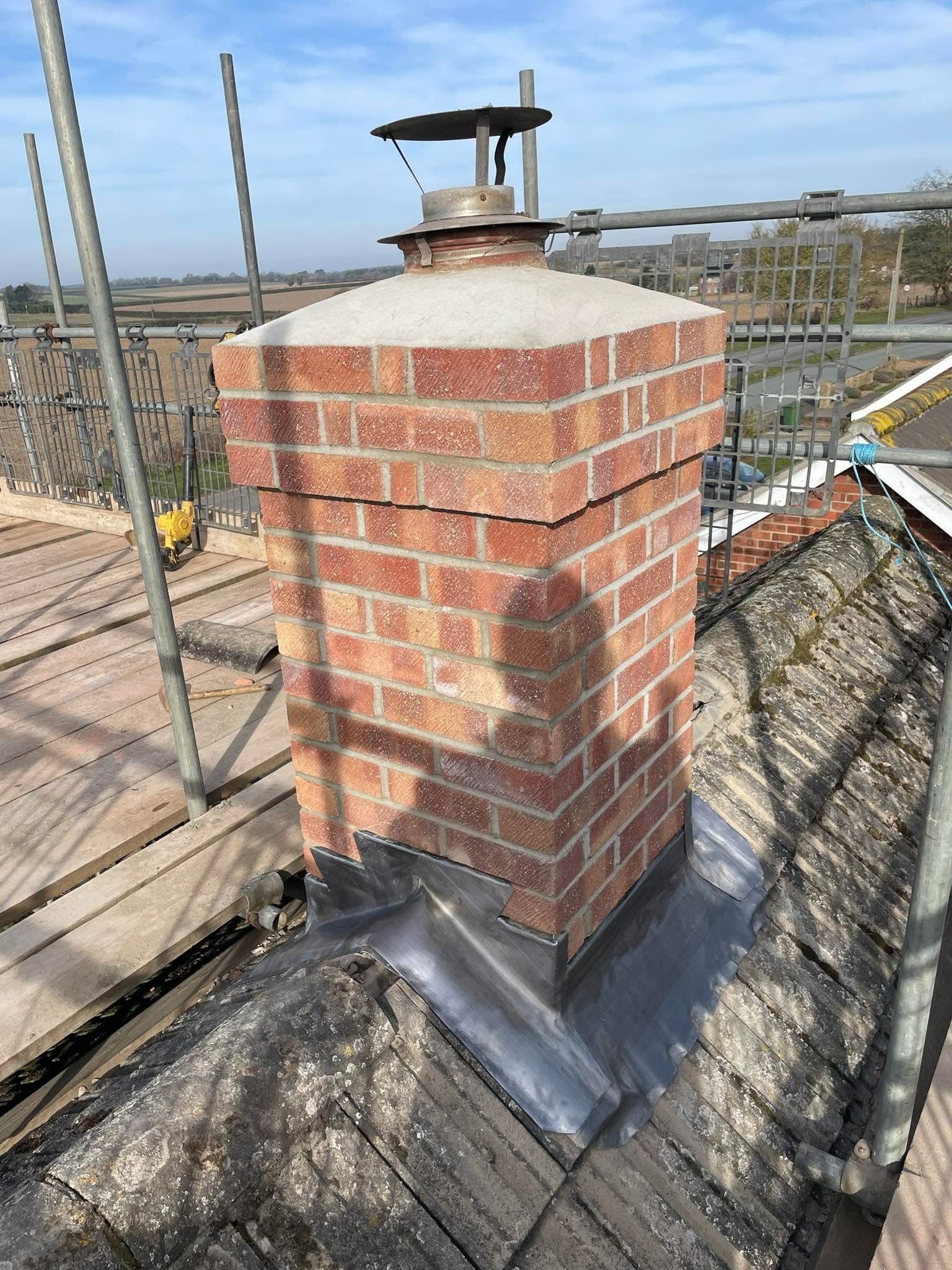 A brick chimney stack on a tiled roof with lead flashing and a metal chimney cowl, surrounded by scaffolding.