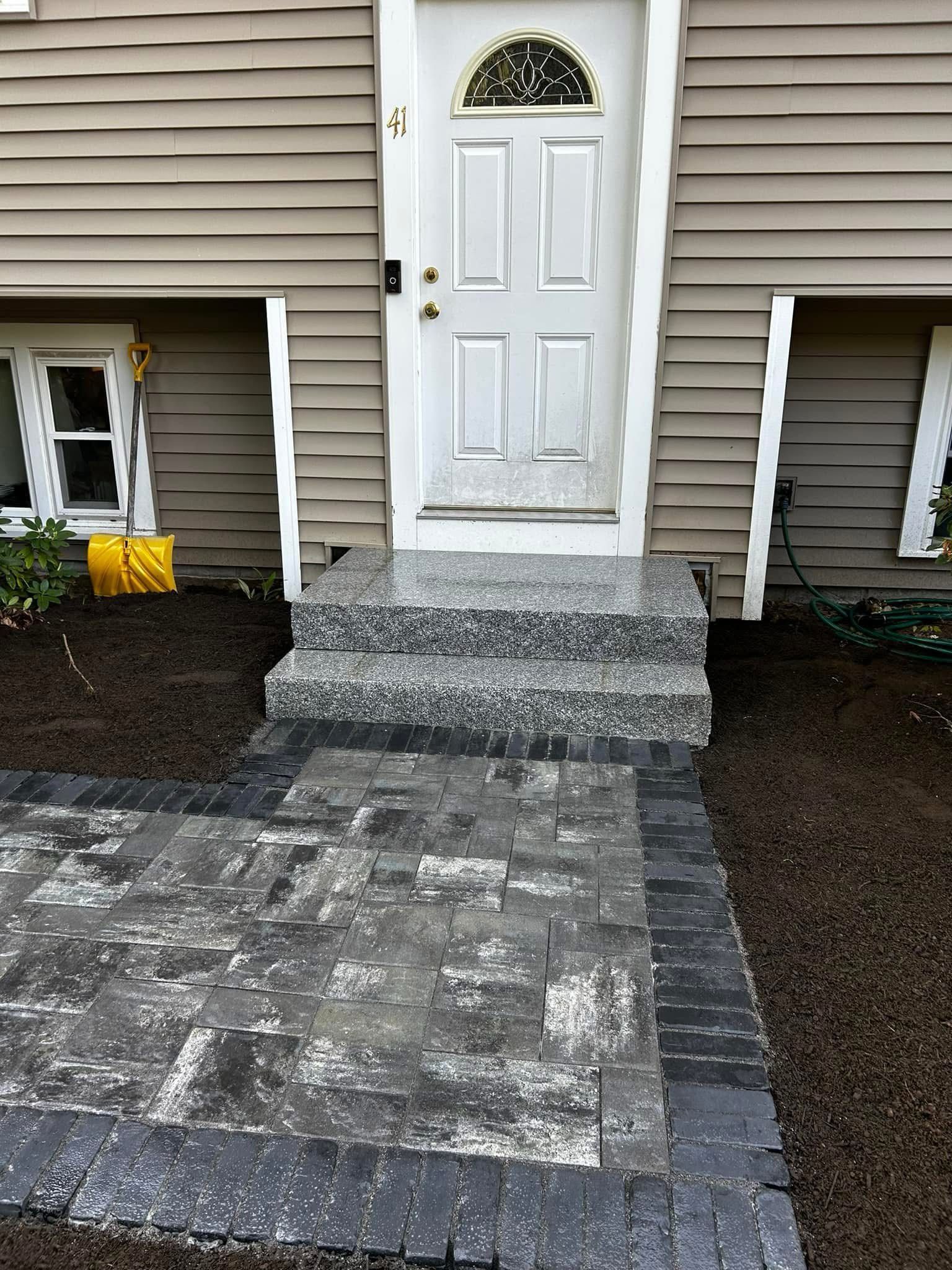 A front entrance with two granite steps leading to a white door, set above a dark paved walkway and garden beds.
