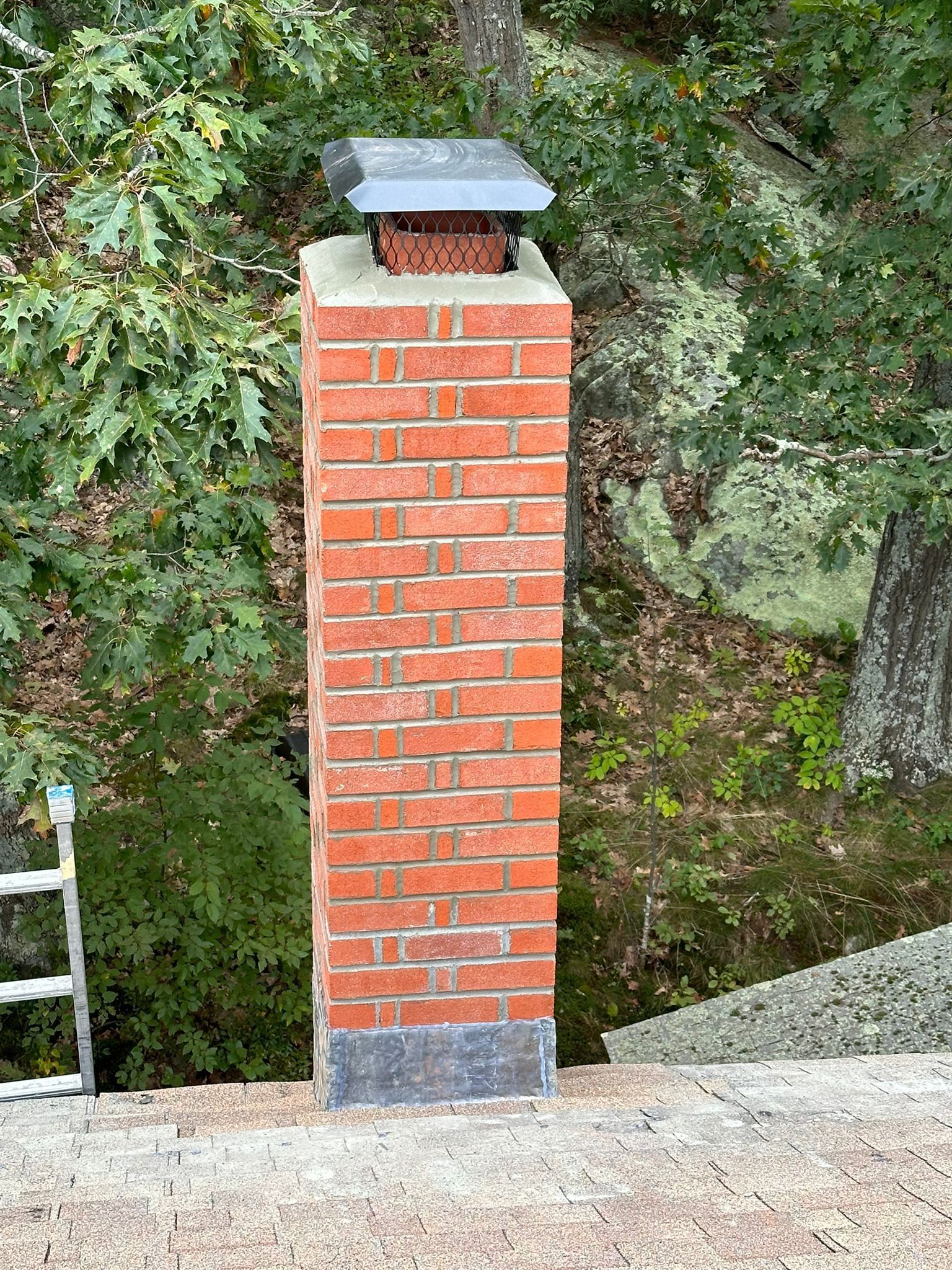 A brick chimney standing on a roof against a background of green trees and rocks.