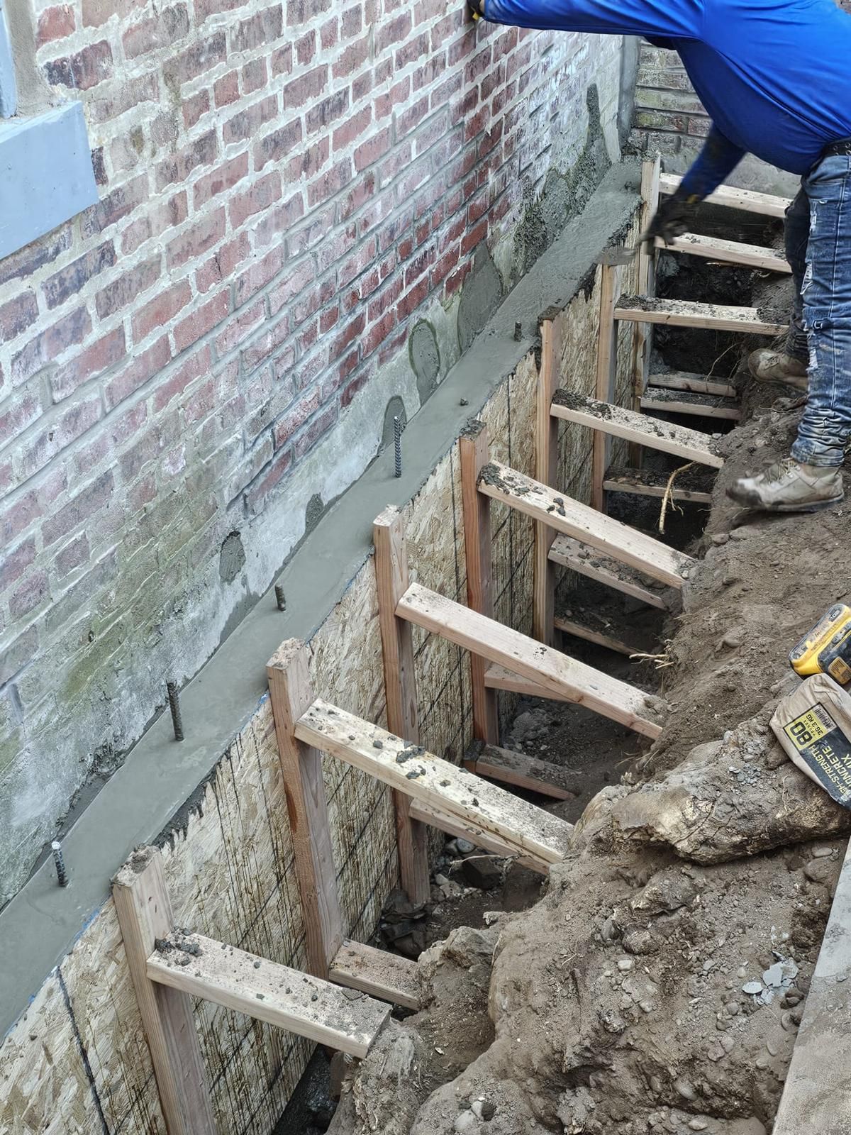 Construction workers pouring a concrete footing against a brick wall, supported by wooden formwork in an excavated trench.