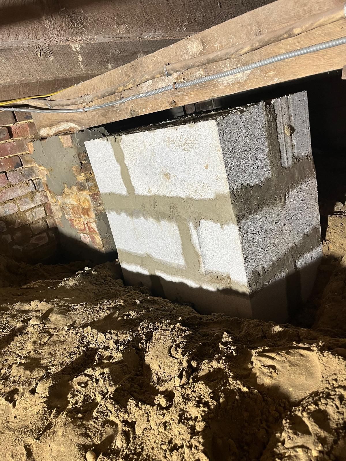 A concrete block pier supports a wooden beam under a building, situated next to an older brick foundation in dirt.