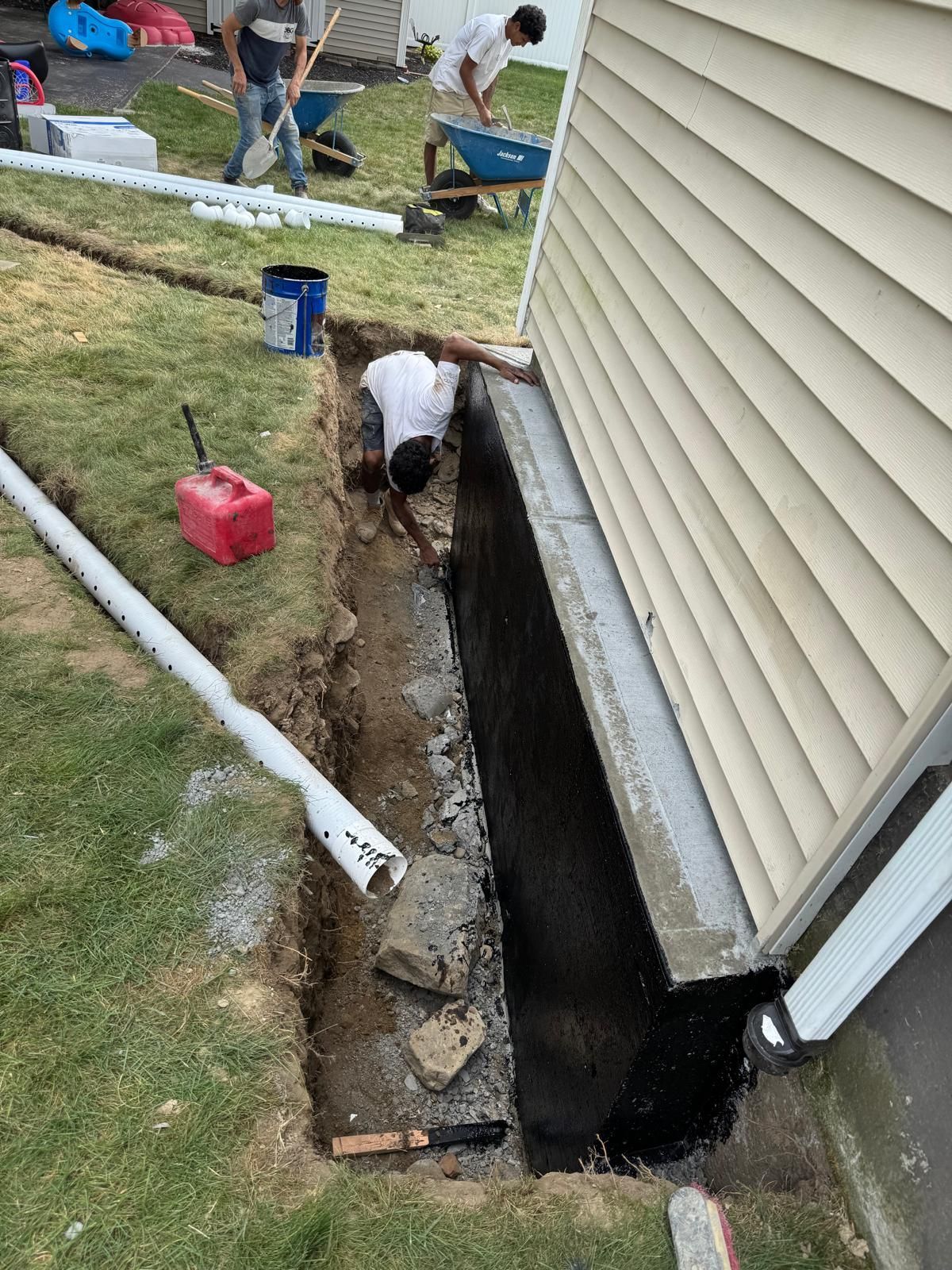 Workers excavate a trench along a home's foundation, applying a dark waterproofing sealant to the concrete wall.