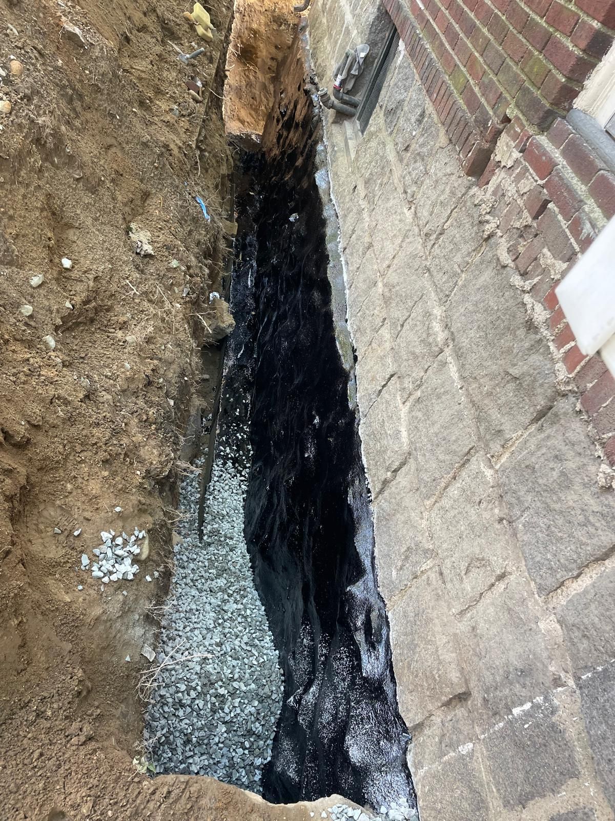 A narrow, excavated trench along a stone foundation wall, coated with black waterproof sealant over a gravel drainage base.