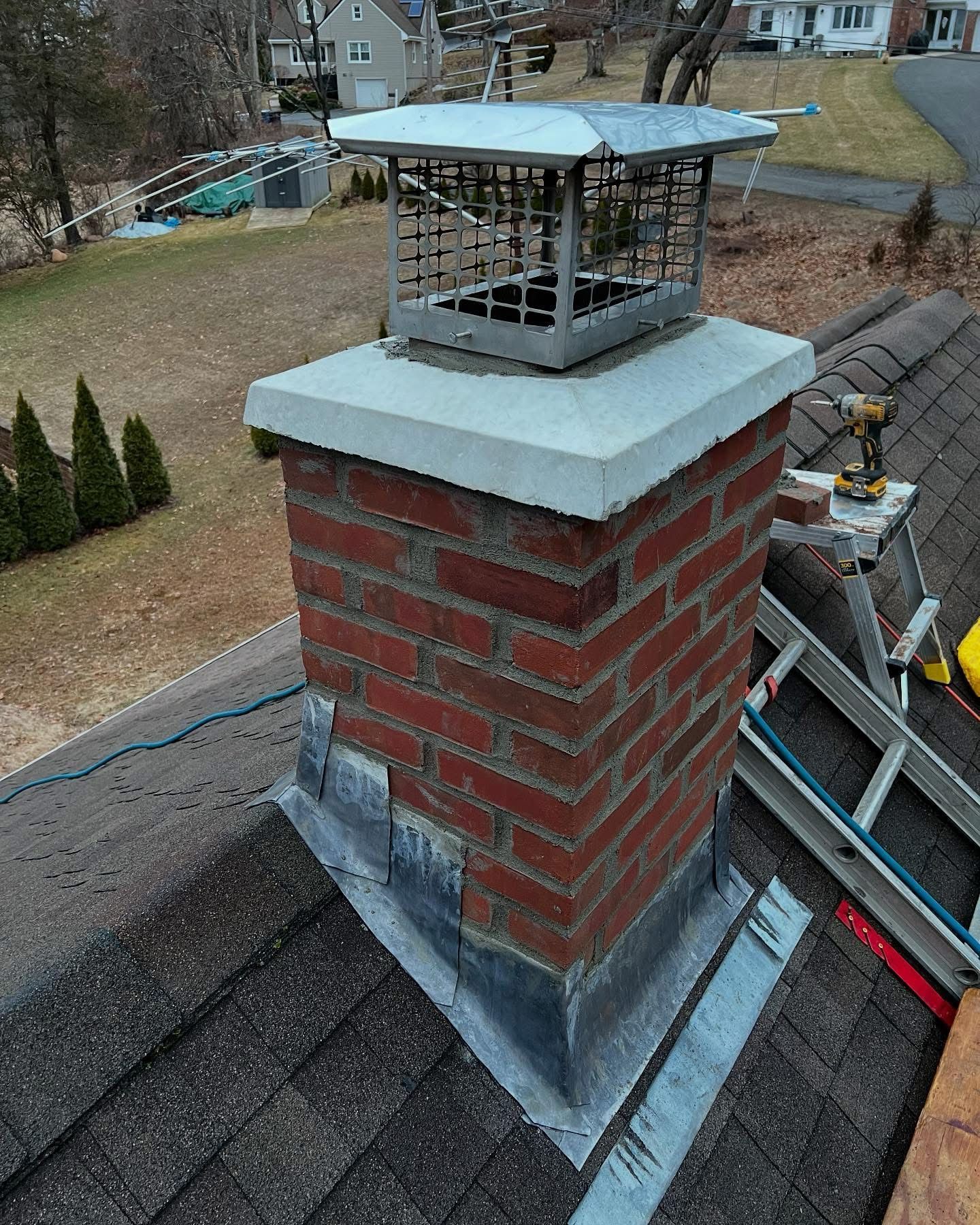 A red brick chimney with a metal cap and flashing on an asphalt shingle roof, with a ladder and tools visible nearby.