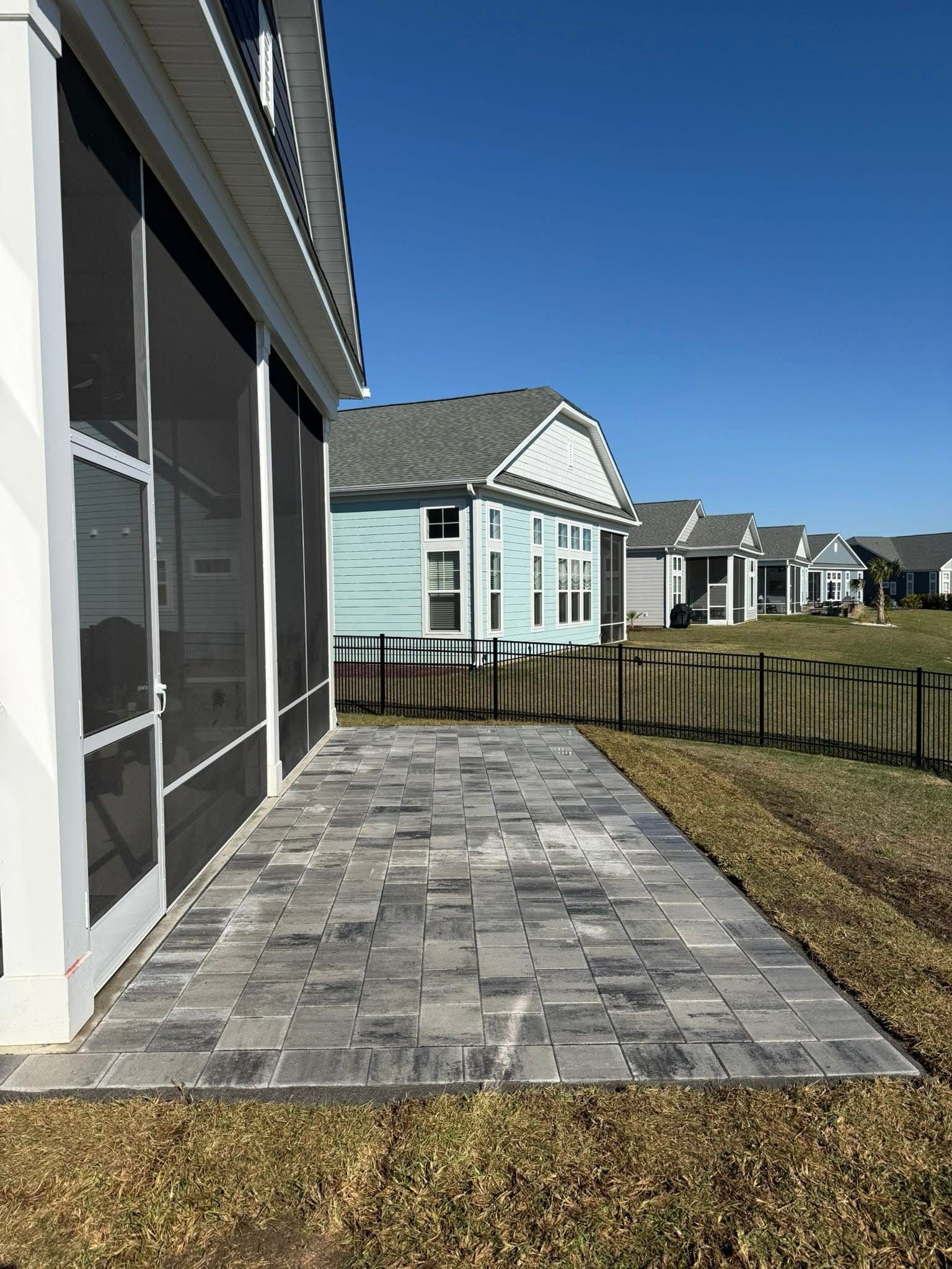 A patio of grey pavers sits beside a screened porch with a blue house and other homes in the background under a blue sky.