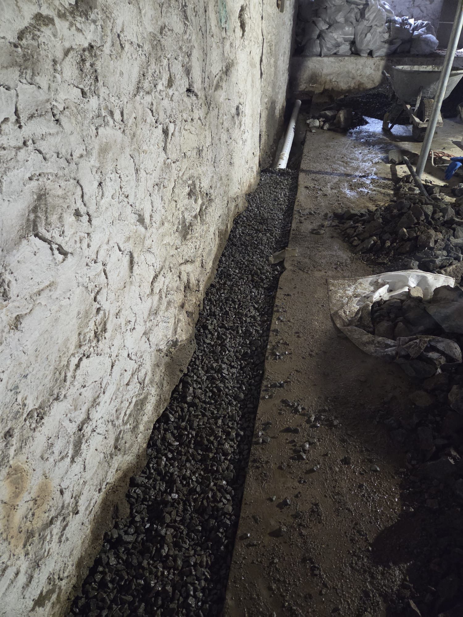 A gravel-filled drainage trench installed along the base of a textured stone basement wall.