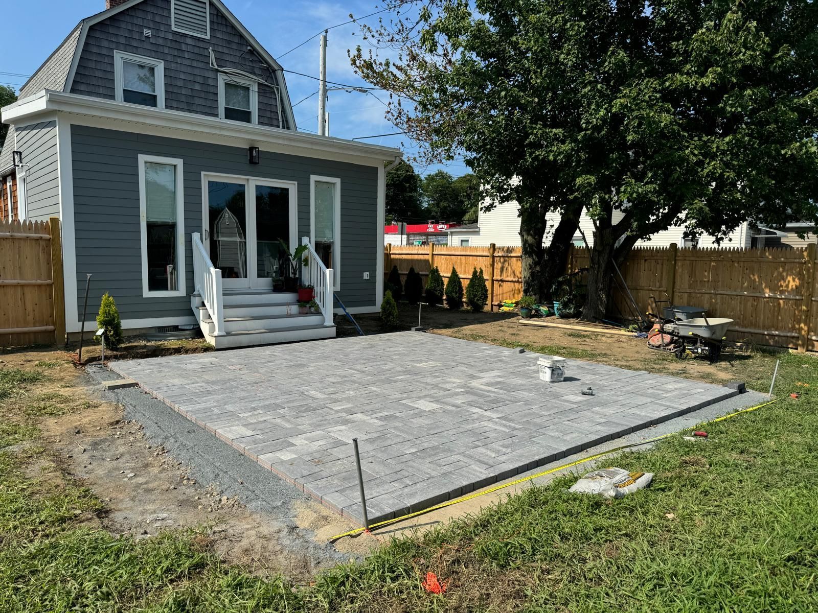 A grey, newly installed paver patio in a backyard sits in front of a house with white stairs leading to glass doors.