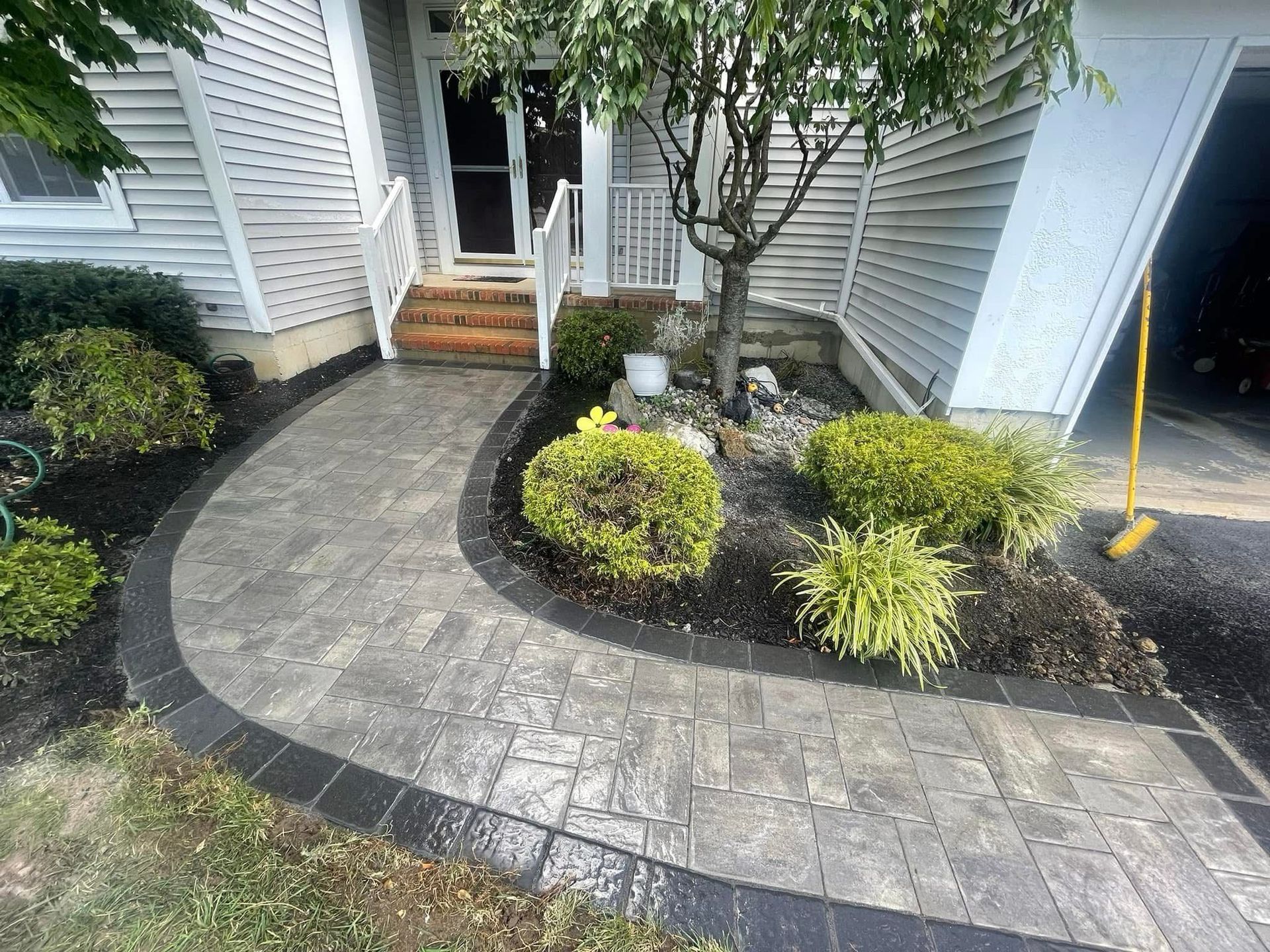 A curved paver walkway with a dark border leads to the front steps of a house with white siding and a small tree.