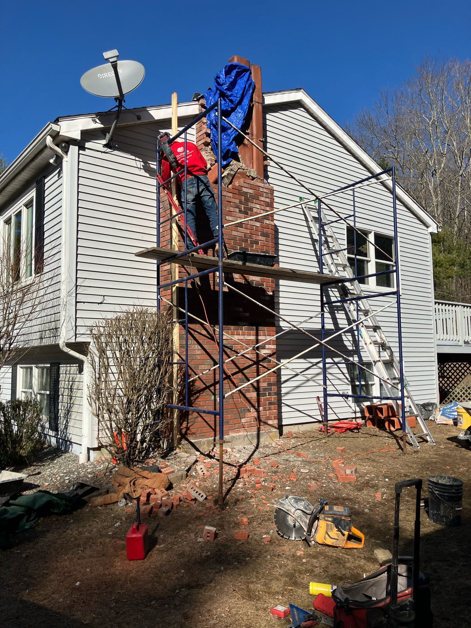 A worker in a red shirt stands on scaffolding, repairing a brick chimney on the side of a house with blue tarp on top.