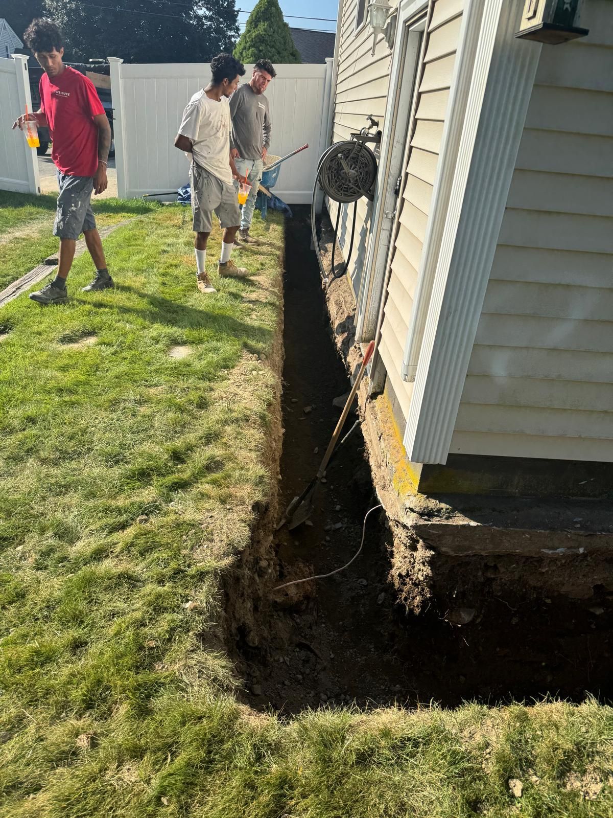Three people stand near a deep trench excavated along the side of a house foundation in a residential yard.