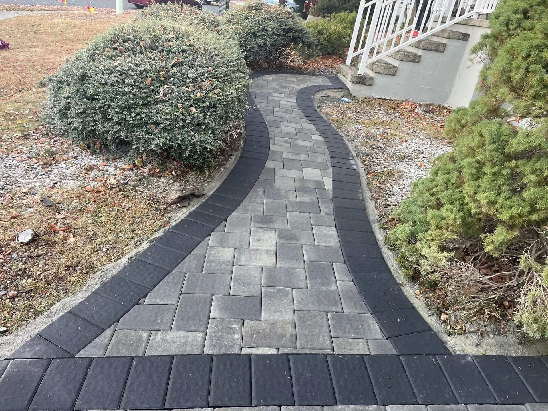 A curving stone walkway with a dark border leads toward a set of steps by a house, surrounded by landscaping bushes.