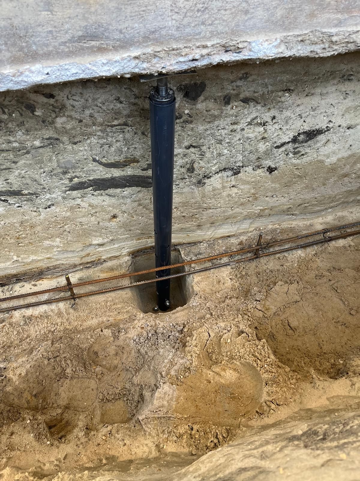A black vertical pile installed beneath an existing concrete foundation in an excavated trench with exposed rebar.
