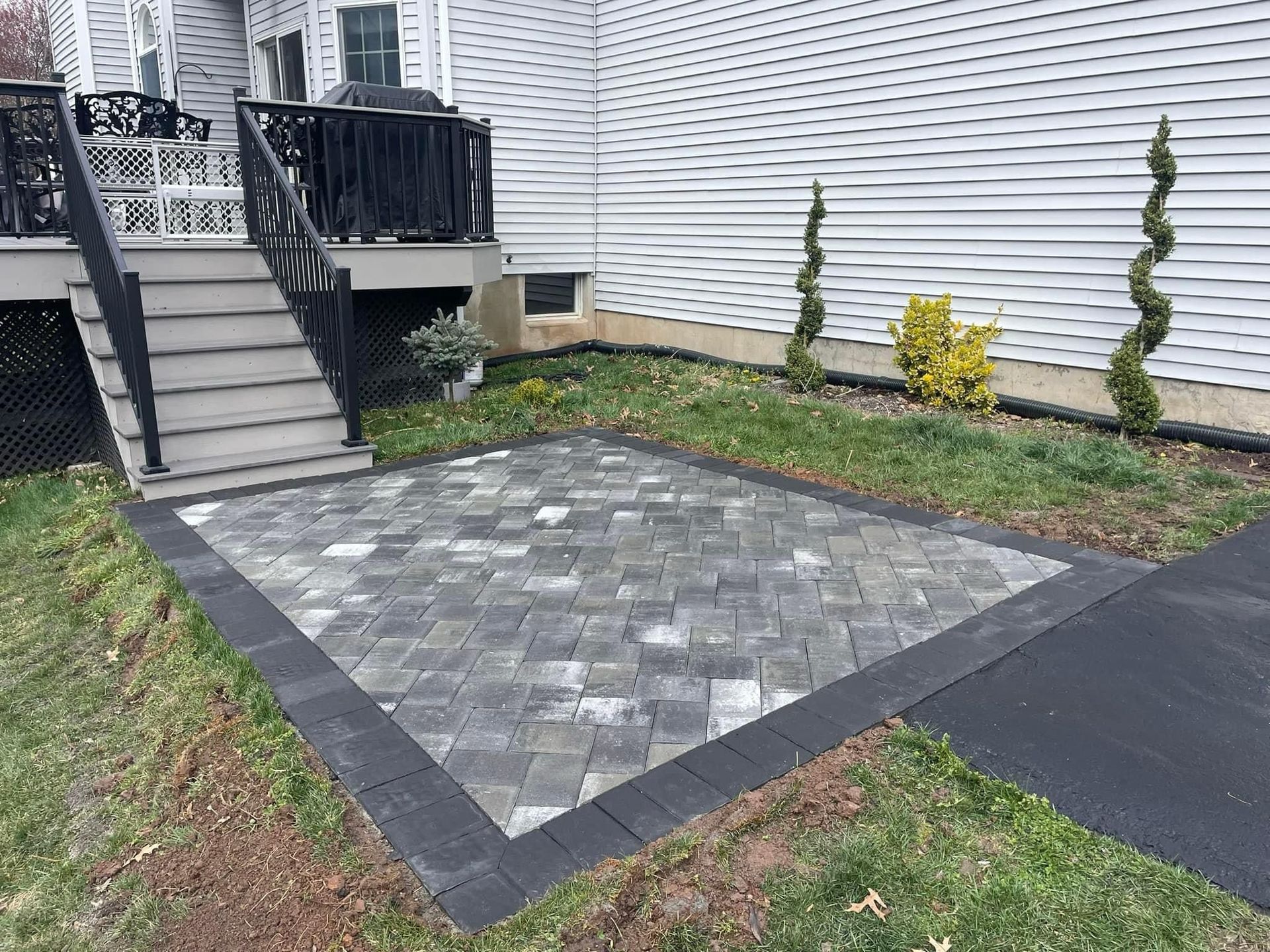A ground-level patio made of grey rectangular pavers with a dark border, located at the base of grey wooden deck stairs.