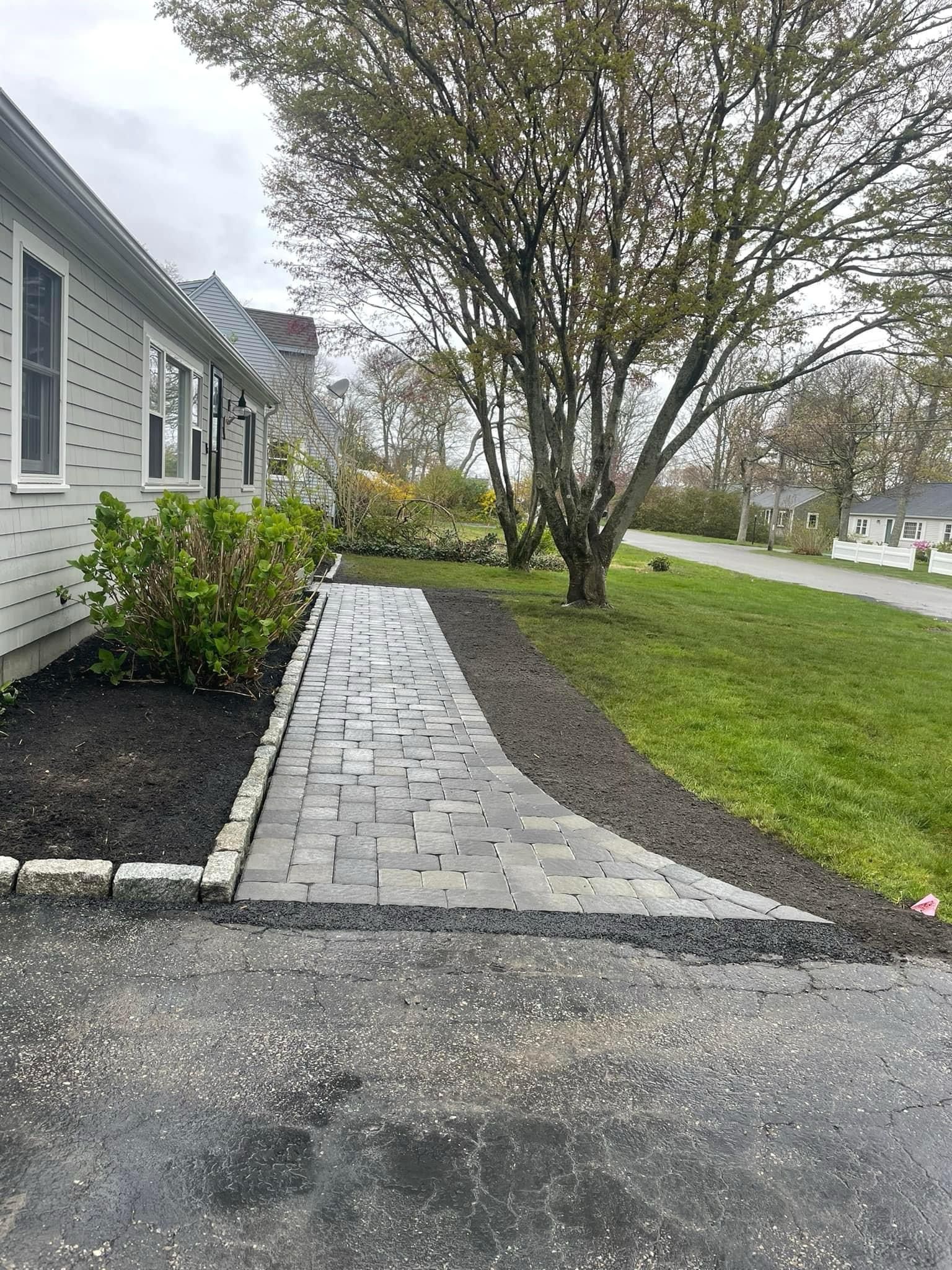 A new gray paver walkway leads from an asphalt driveway to a house, flanked by black mulch and a large tree.