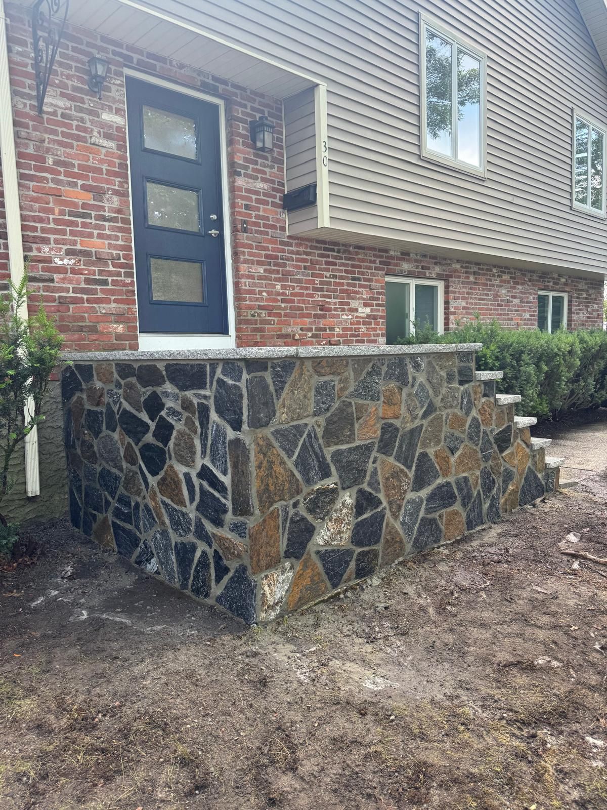 Front door of a house with siding and brick, featuring a newly constructed stone porch with a multi-colored flagstone finish.