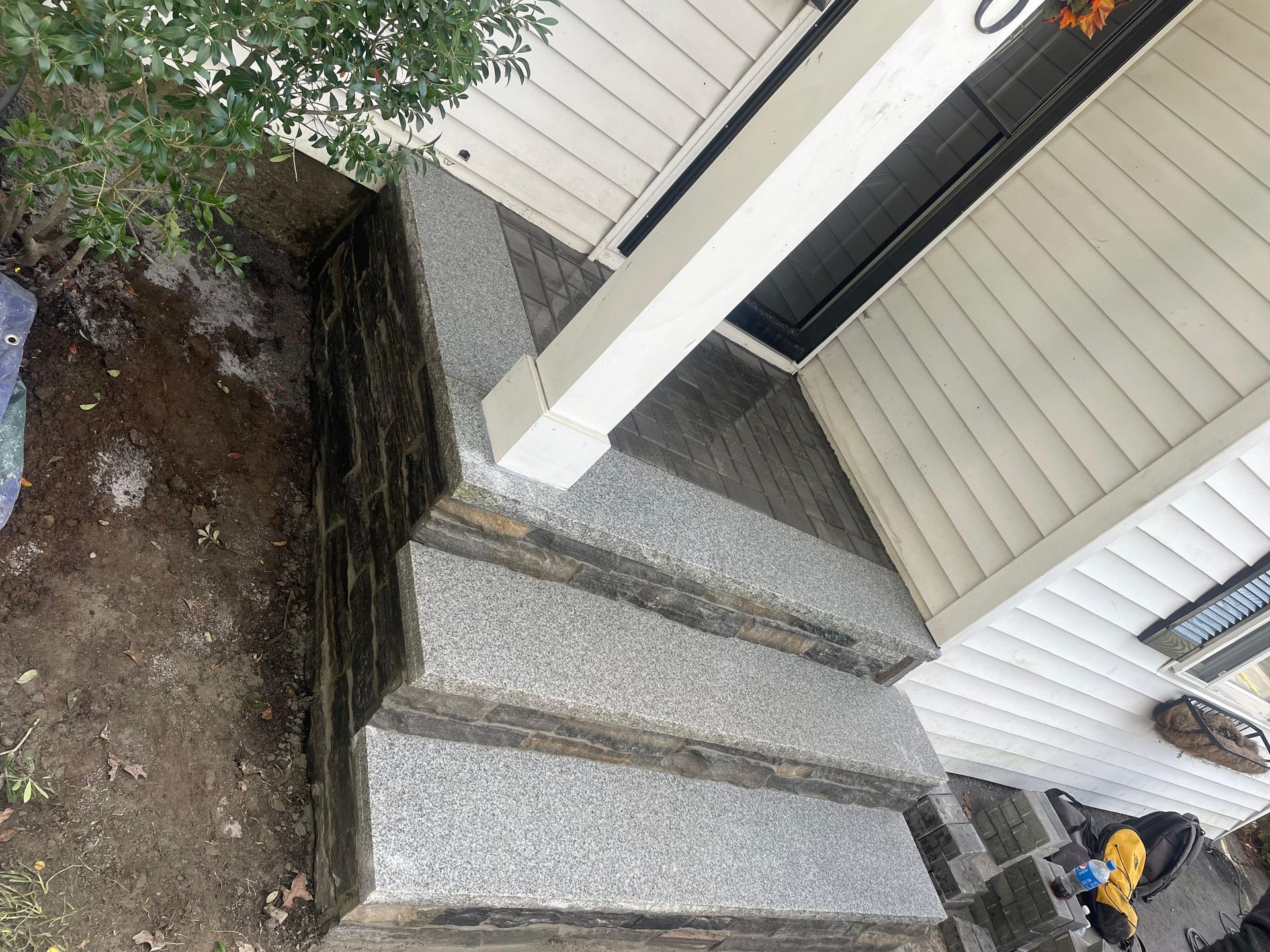 A set of three stone steps with gray granite treads leading to a house entrance with beige siding and a white column.