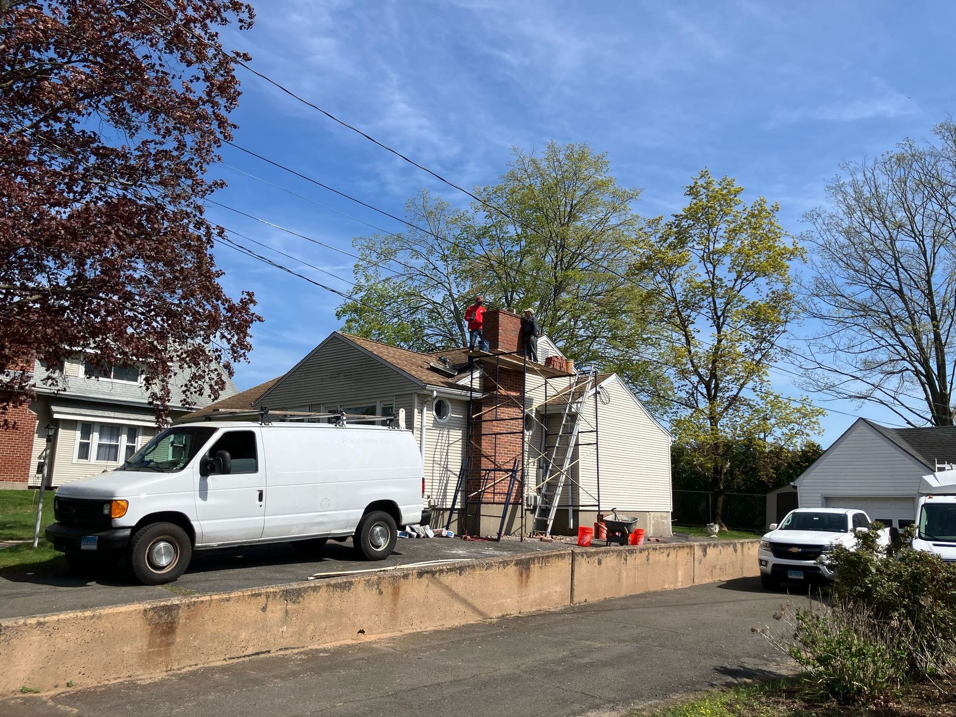 Workers use scaffolding to perform repairs on a brick chimney attached to a light-colored house with a van parked nearby.