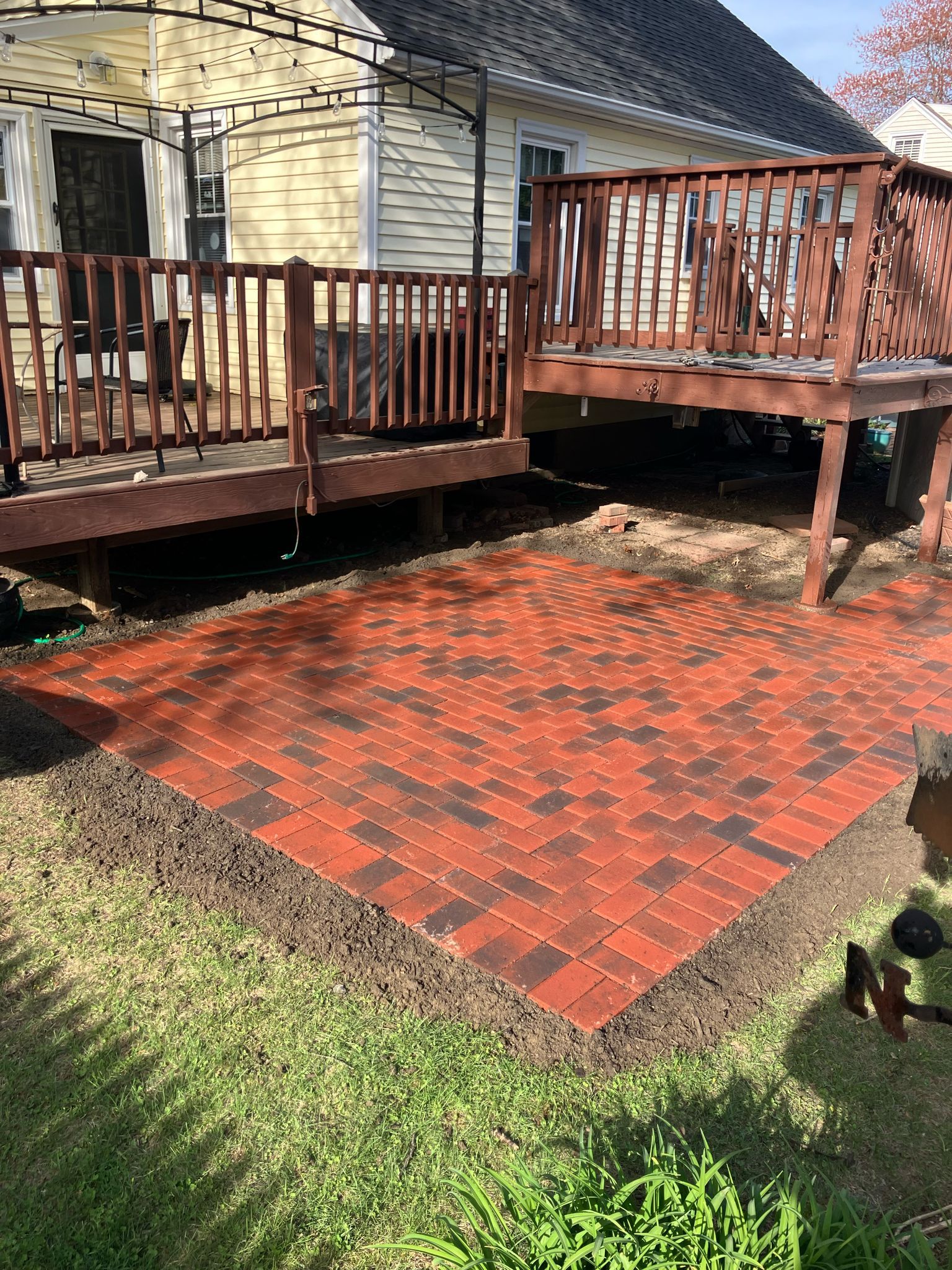 A rectangular brick patio made of red and brown pavers sits in a yard in front of a wooden deck and a yellow house.
