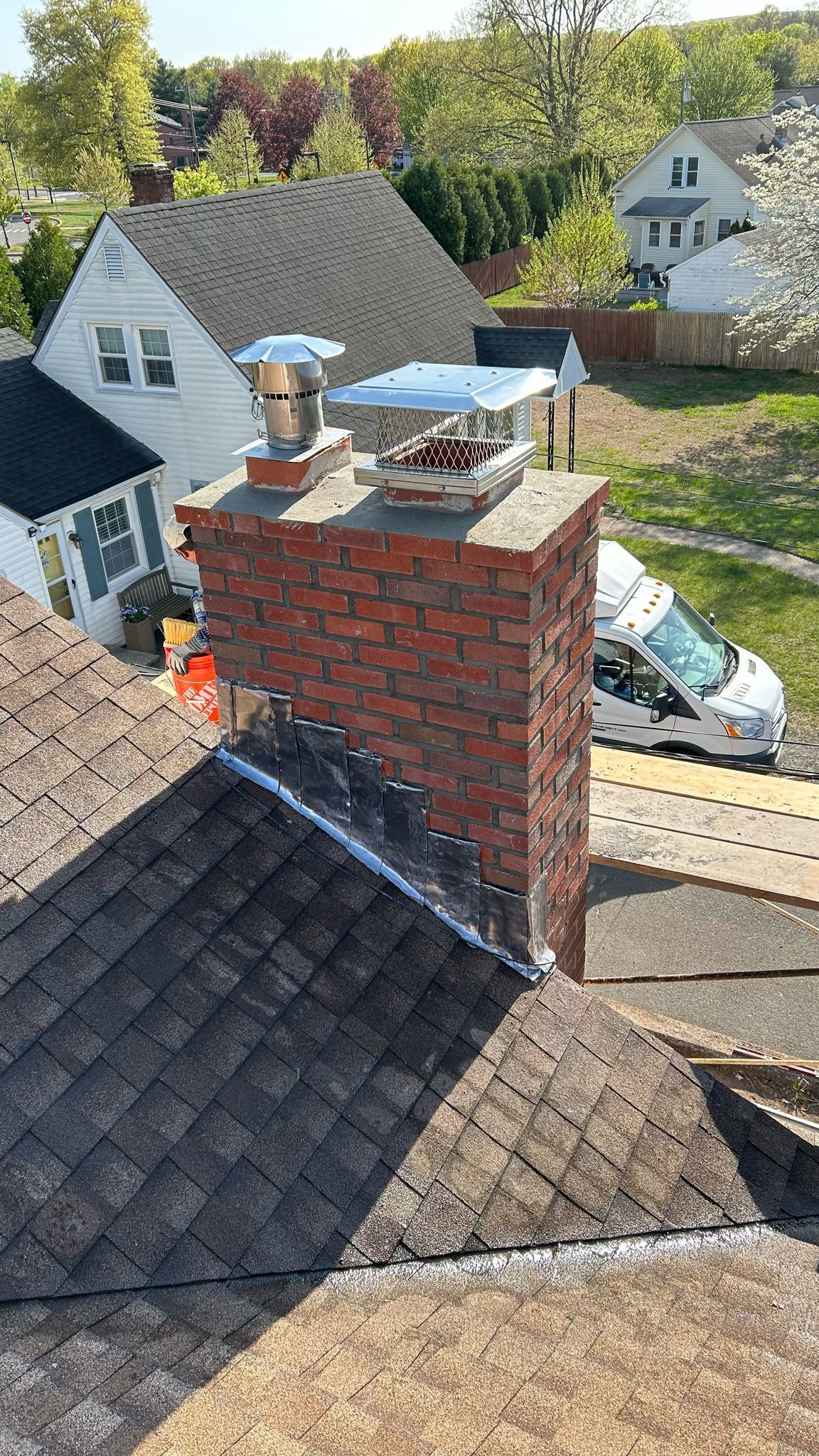 High-angle view of a red brick chimney on a shingled roof with two metal flue caps, next to a white house.